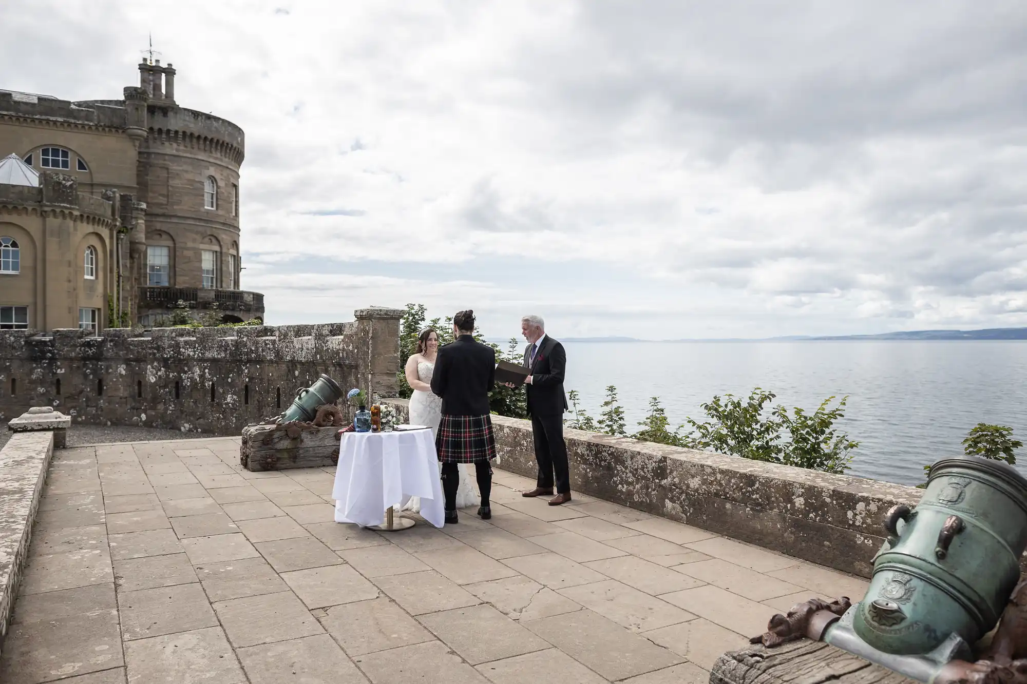 A couple stands with an officiant near a table on a stone terrace overlooking a body of water with an old castle building in the background.