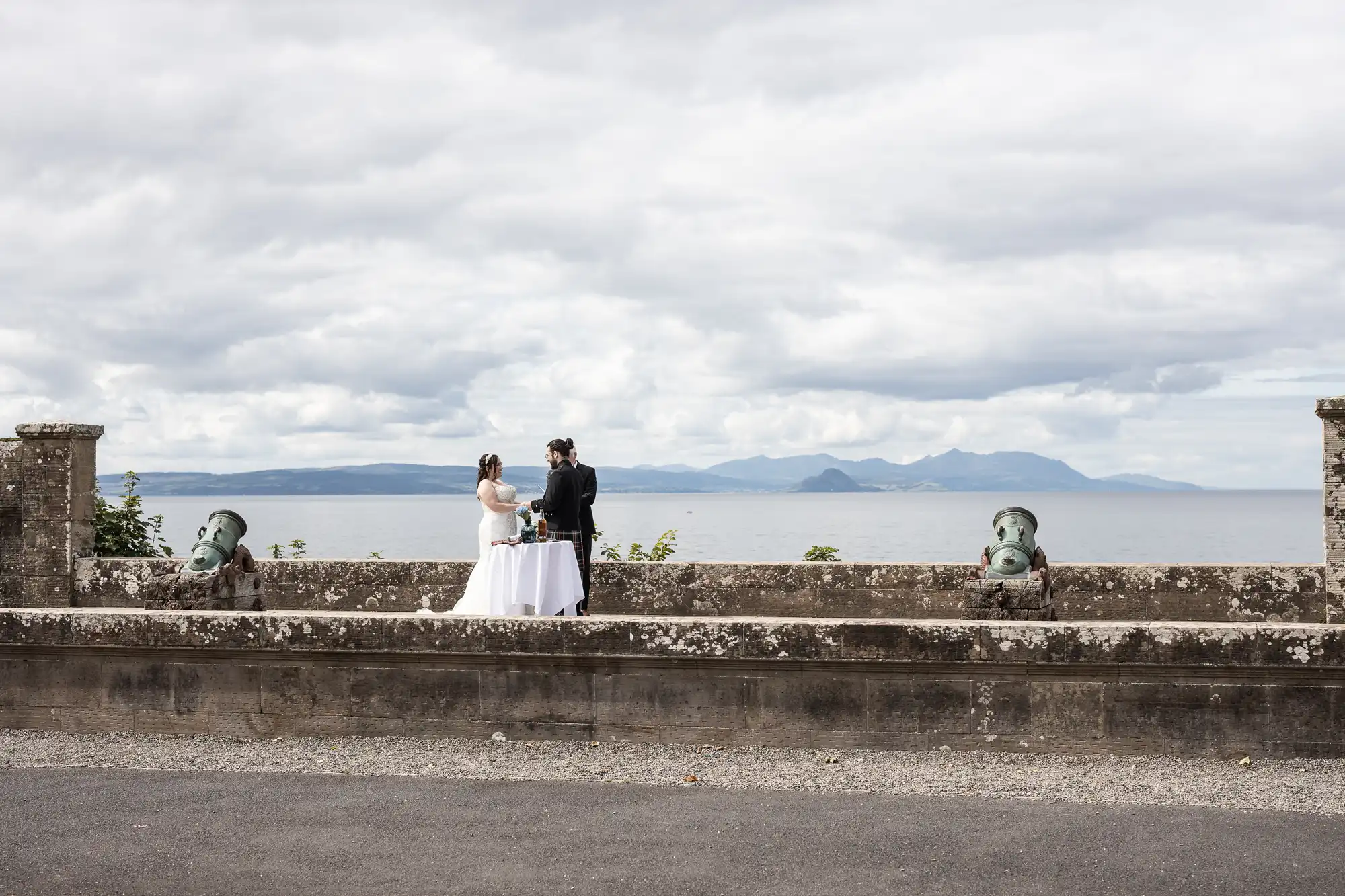 A couple stands at an outdoor altar set between two cannons on a stone platform, with mountains and an ocean in the background under a cloudy sky.