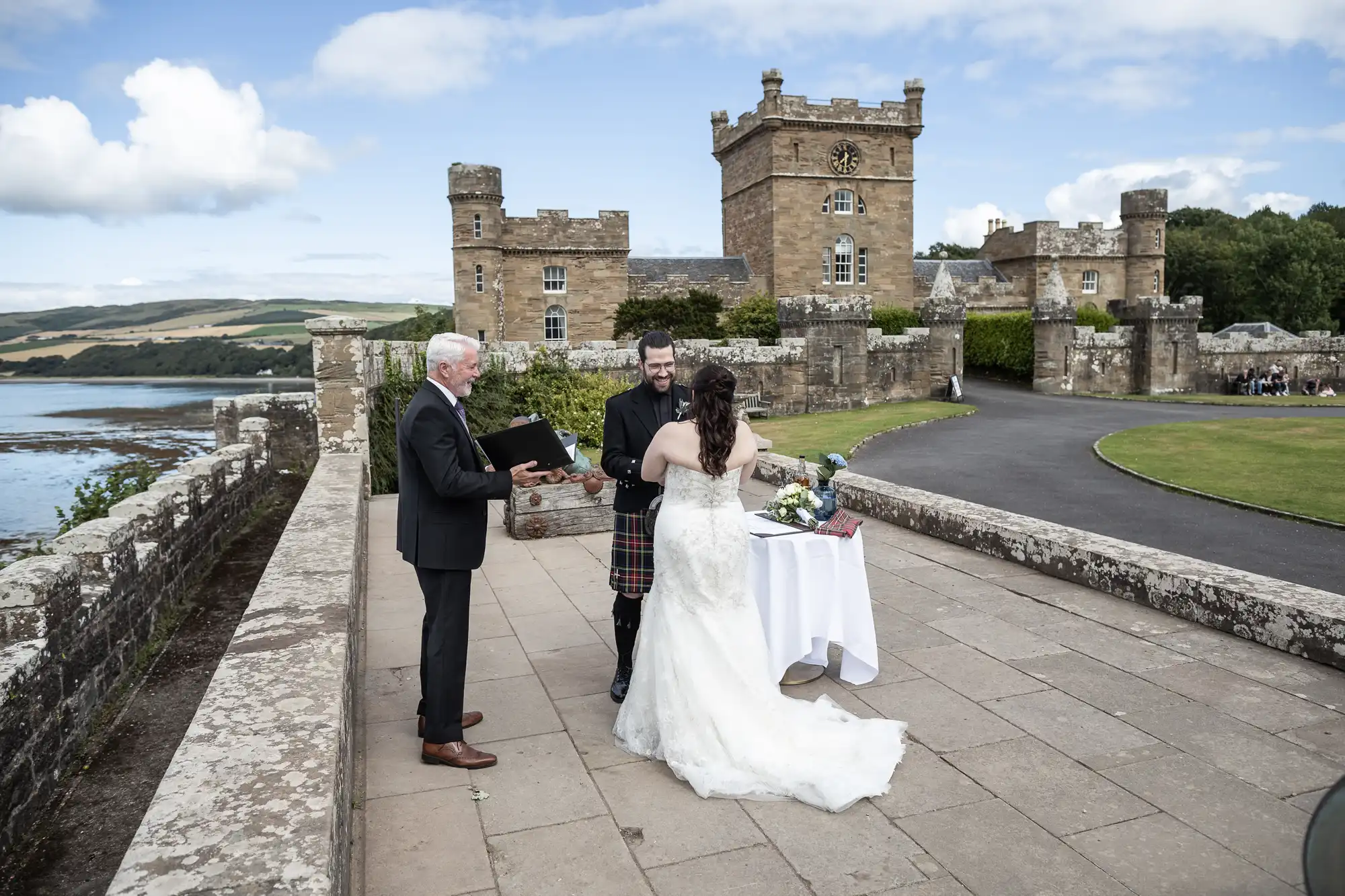 A couple stands before an officiant during an outdoor wedding ceremony at a stone castle, with the bride in a white dress and the groom in Scottish attire.