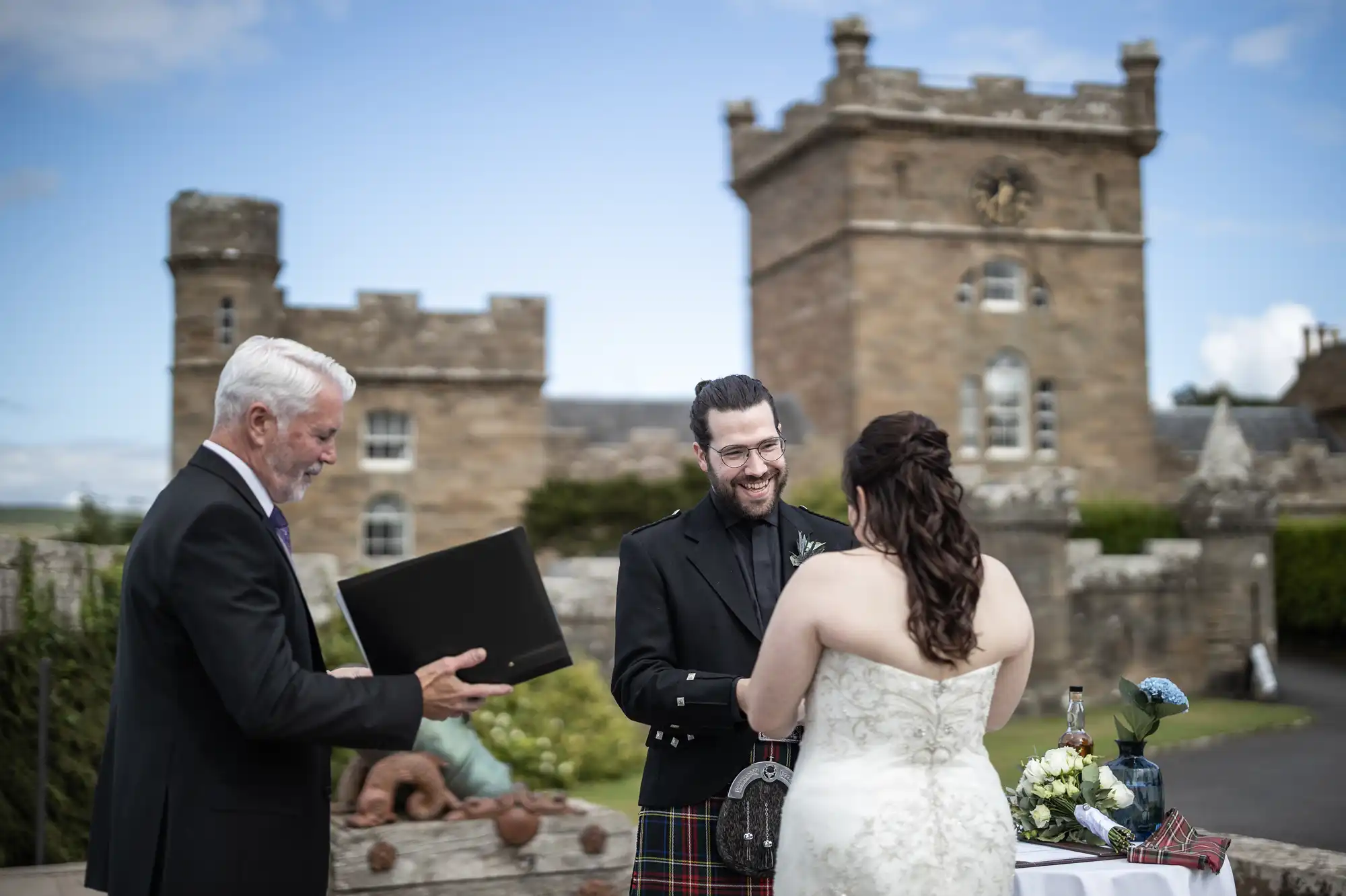 A couple stands before an officiant during an outdoor wedding ceremony at a historic stone castle.