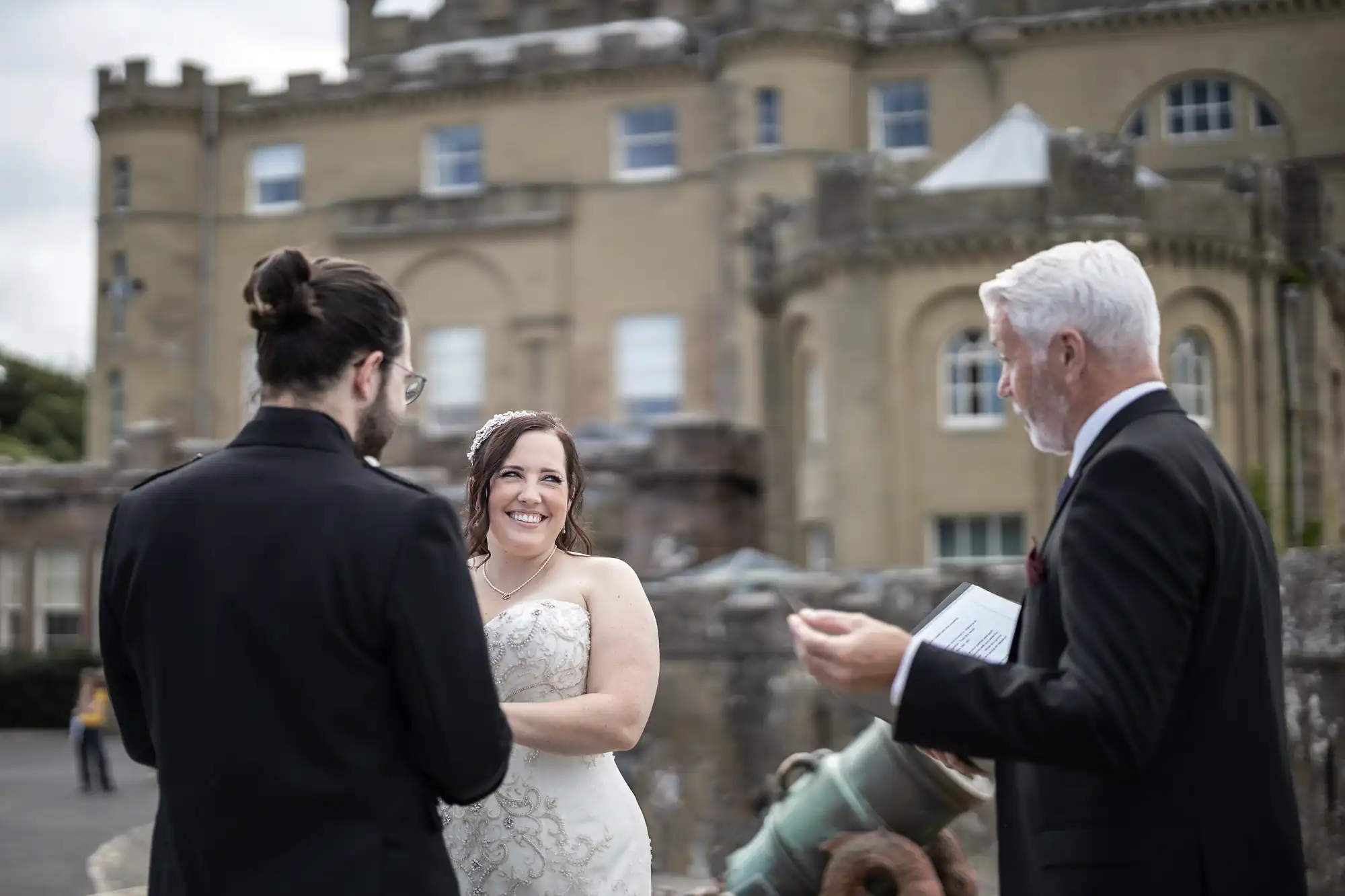 A bride smiles at her groom during an outdoor wedding ceremony, with an officiant speaking beside them. They stand in front of a historic building.