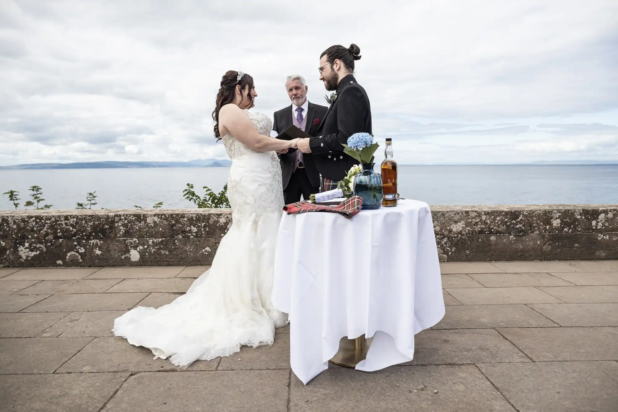 A couple stands facing each other holding hands, with a person in a suit between them officiating their wedding ceremony by the sea. A table with a white cloth, bottle, and items sits nearby.