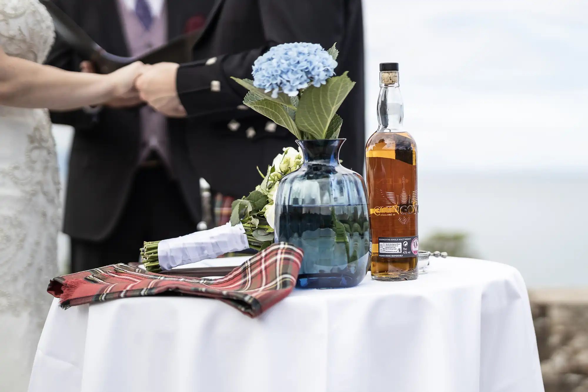 A table with a blue vase holding blue flowers, a bottle of whiskey, and a red plaid cloth in front of a couple holding hands.