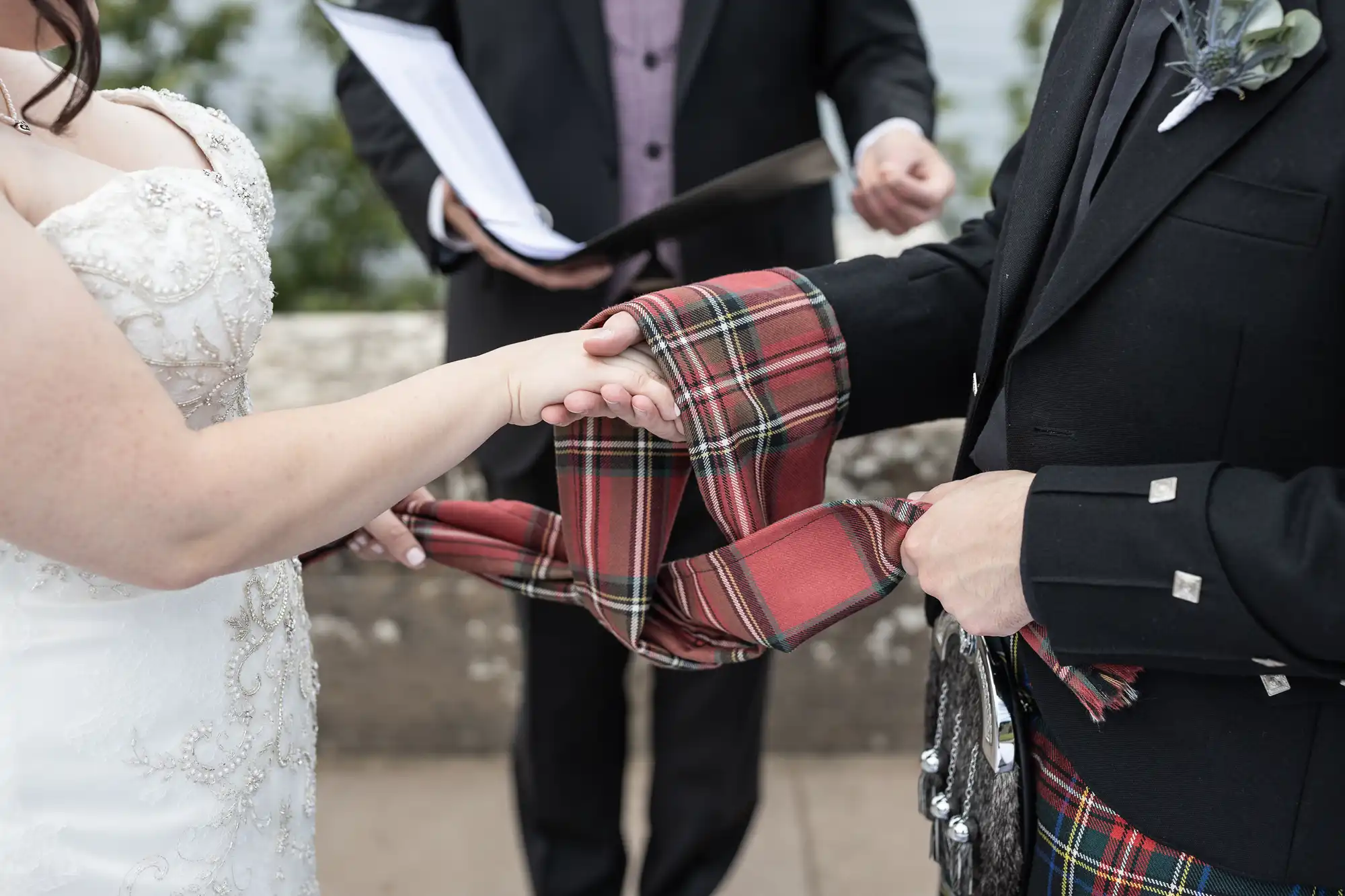 A couple participates in a handfasting ceremony, where their hands are bound with a red tartan cloth held by an officiant dressed in a dark suit.