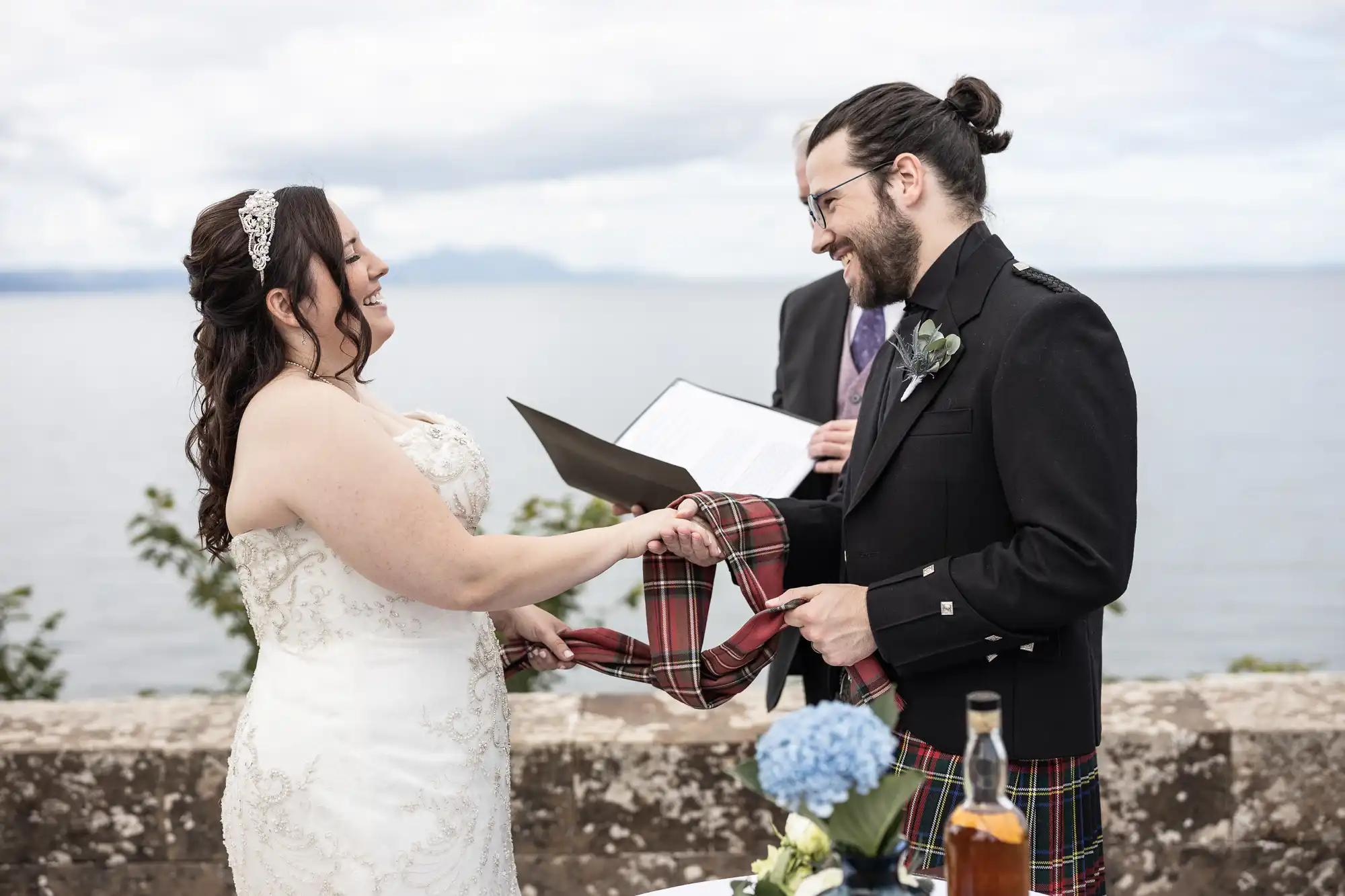 A bride and groom, both smiling, participate in a handfasting ceremony with a tartan cloth by the seaside. The officiant stands nearby holding a book.