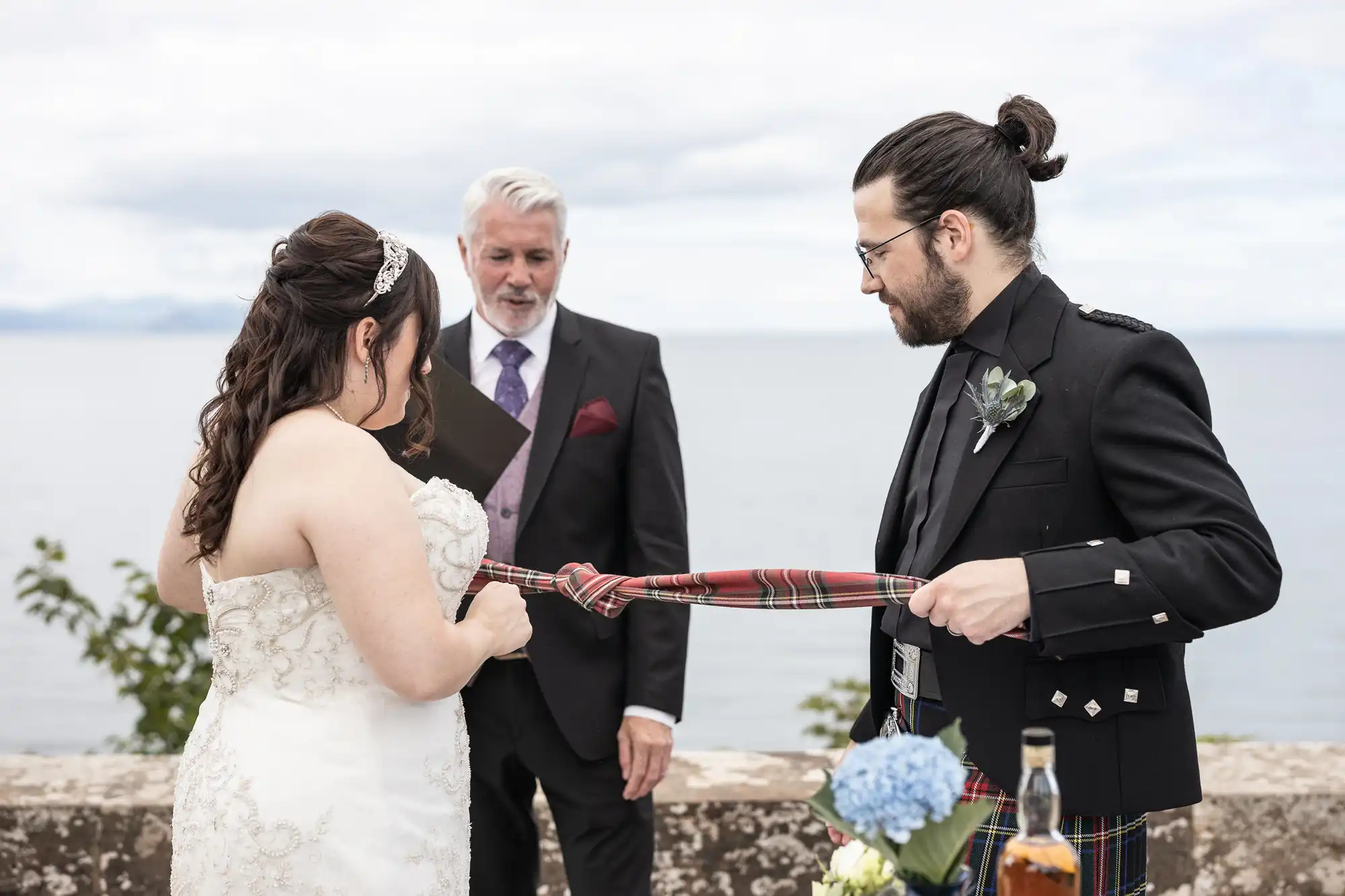 A bride and groom participate in a handfasting ceremony while a man in a suit officiates, set against a scenic ocean backdrop.