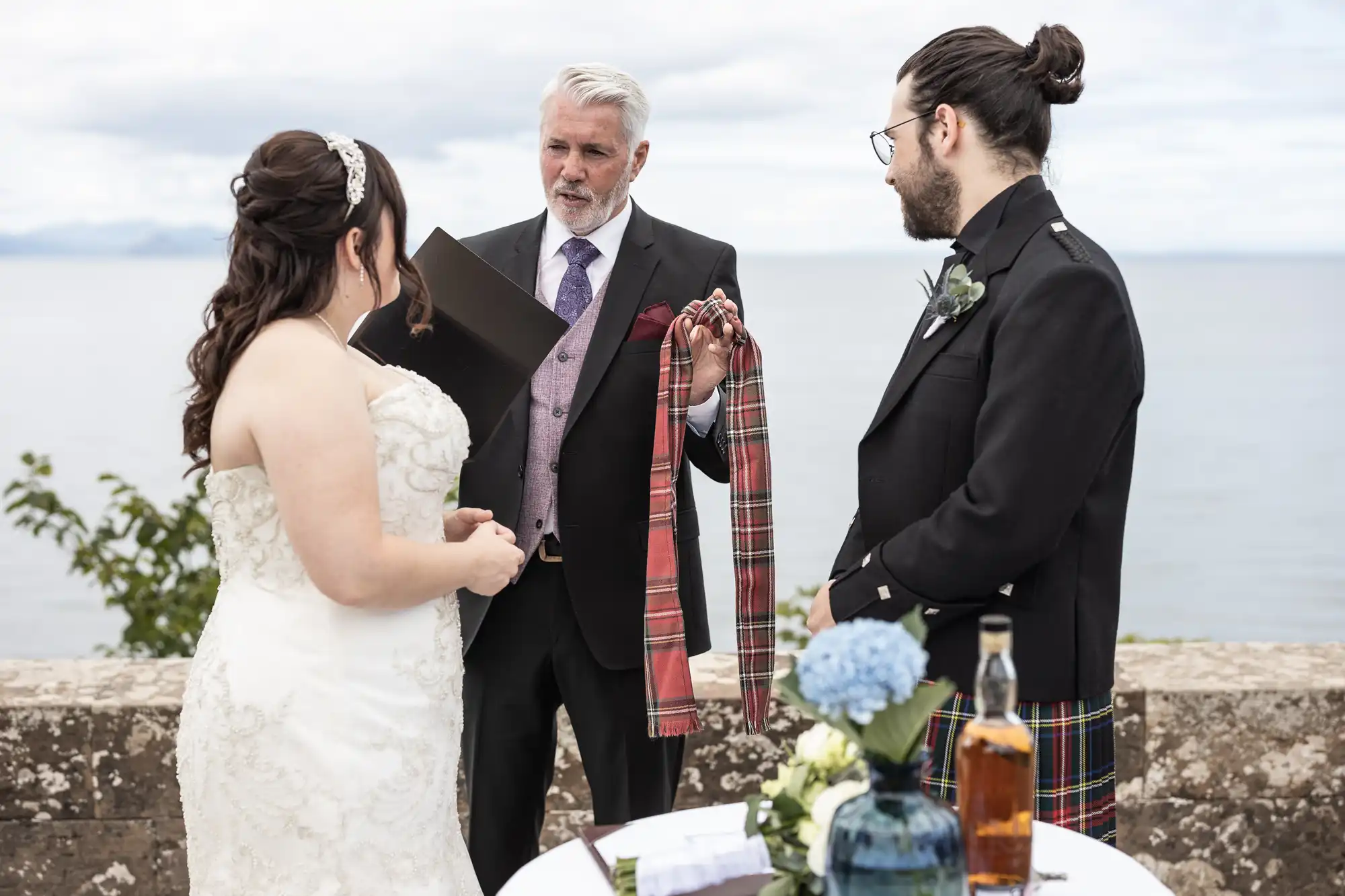 A wedding ceremony by the sea, featuring a couple and an officiant holding a tartan cloth. A table with a bottle and flowers is in the foreground.