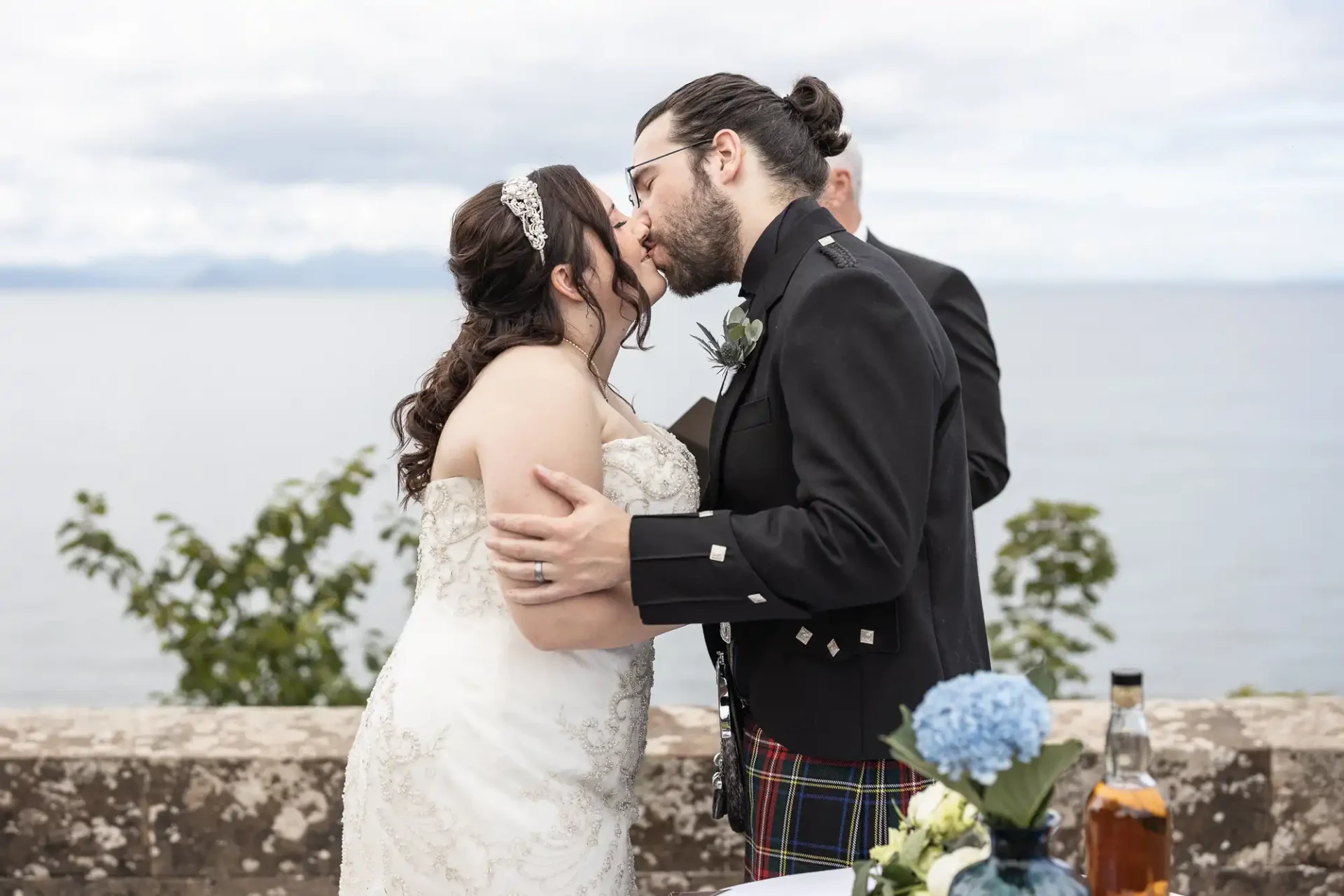 A bride and groom share a kiss during an outdoor wedding ceremony. A scenic body of water and cloudy sky are visible in the background.