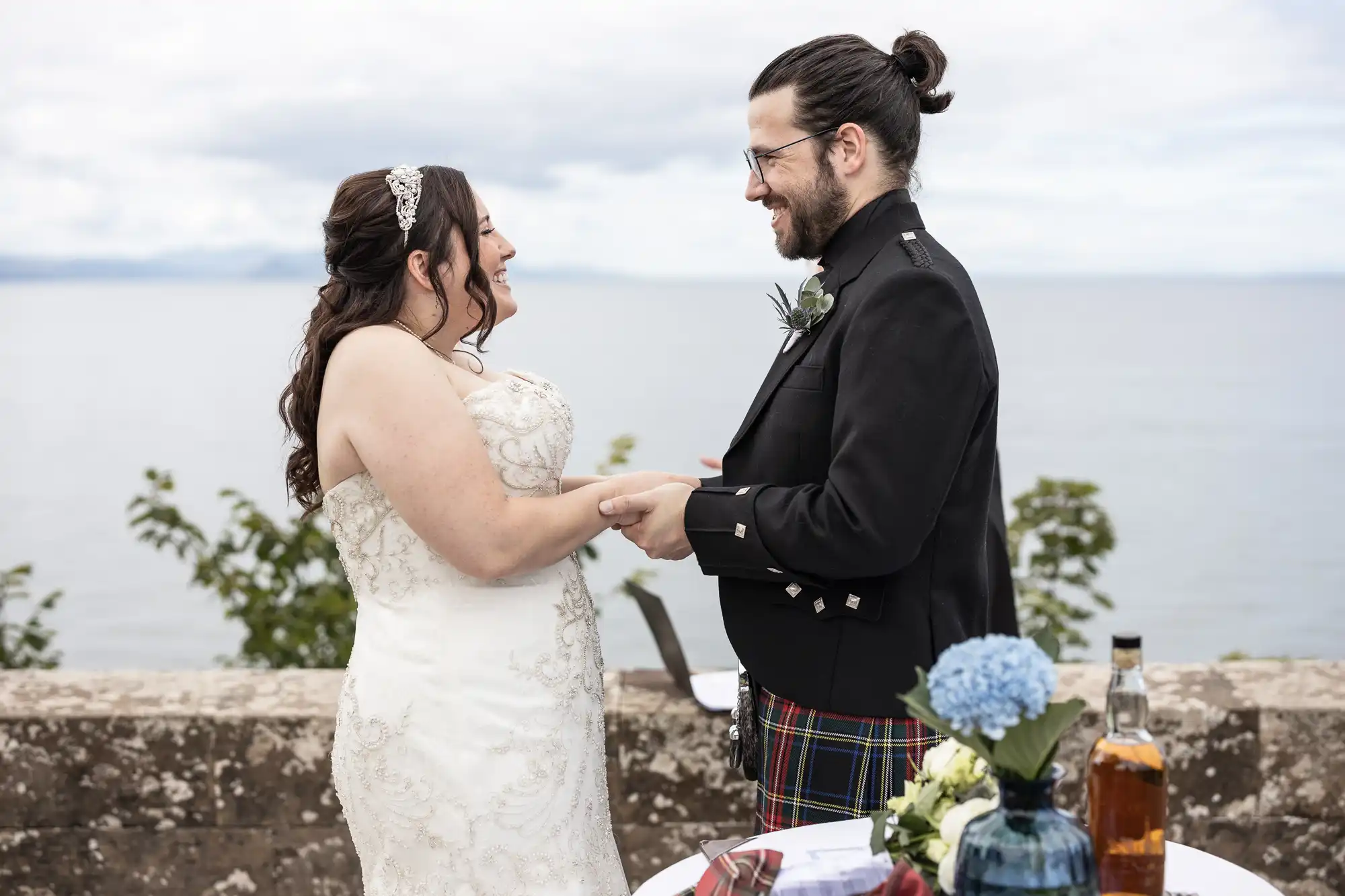 A couple holding hands and smiling at each other during a ceremony. They stand outdoors near a stone wall with a scenic ocean view in the background. A table with decorative items is in the foreground.