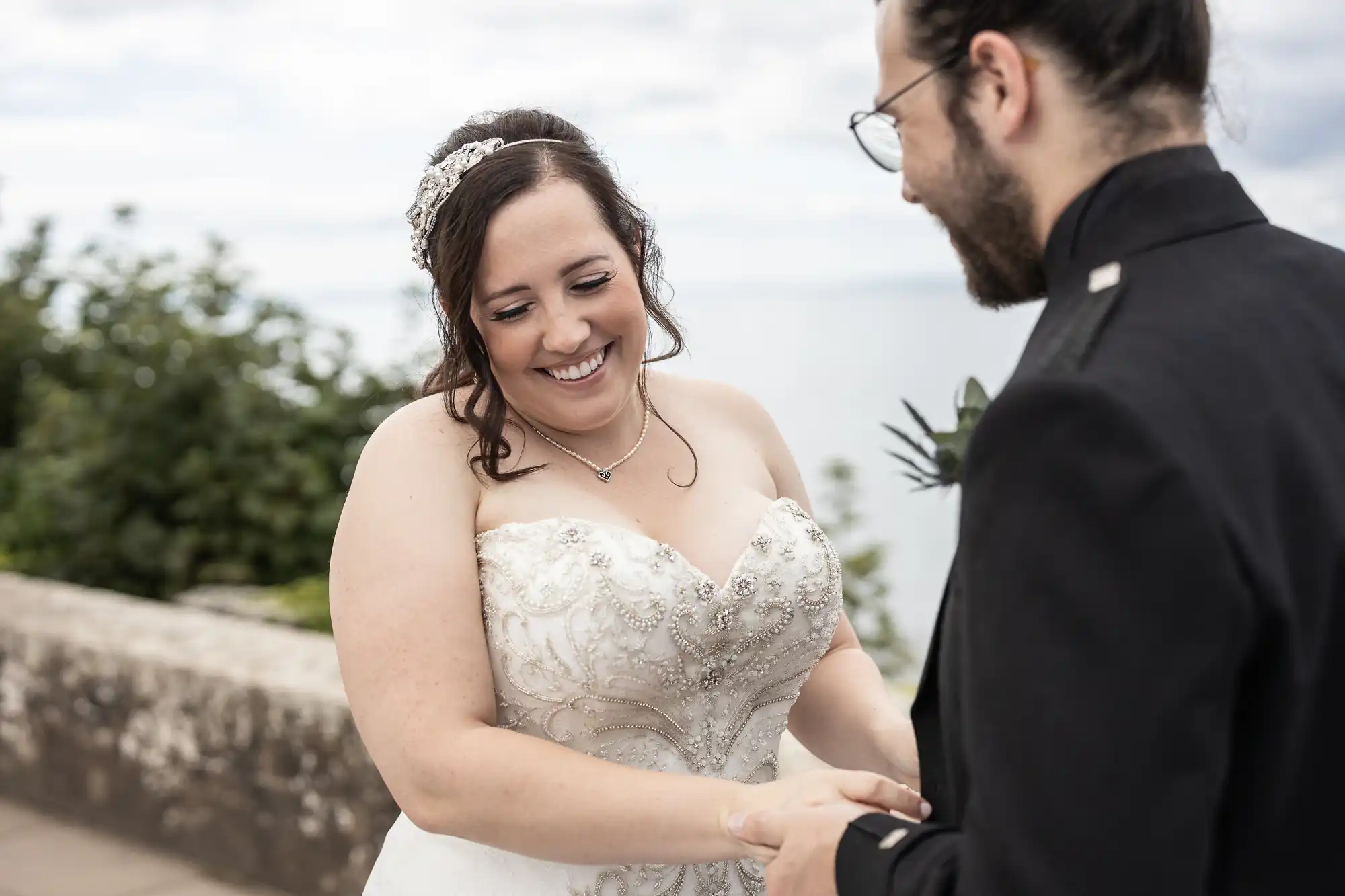 A bride and groom share a moment, smiling at each other while holding hands outdoors, with greenery and the sky in the background.