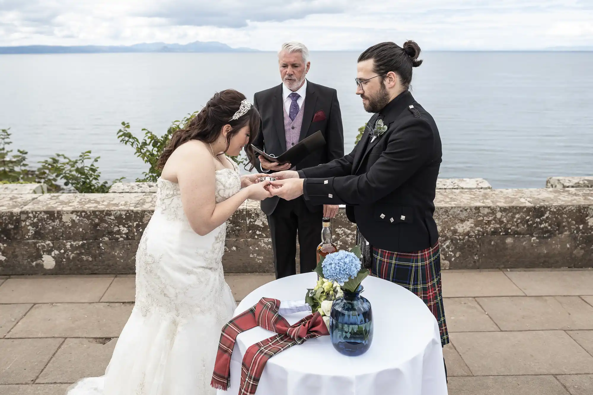 A bride and groom exchange rings in an outdoor wedding ceremony by a body of water, with an officiant standing between them. A small table with a vase of blue flowers and tartan fabric is in the foreground.