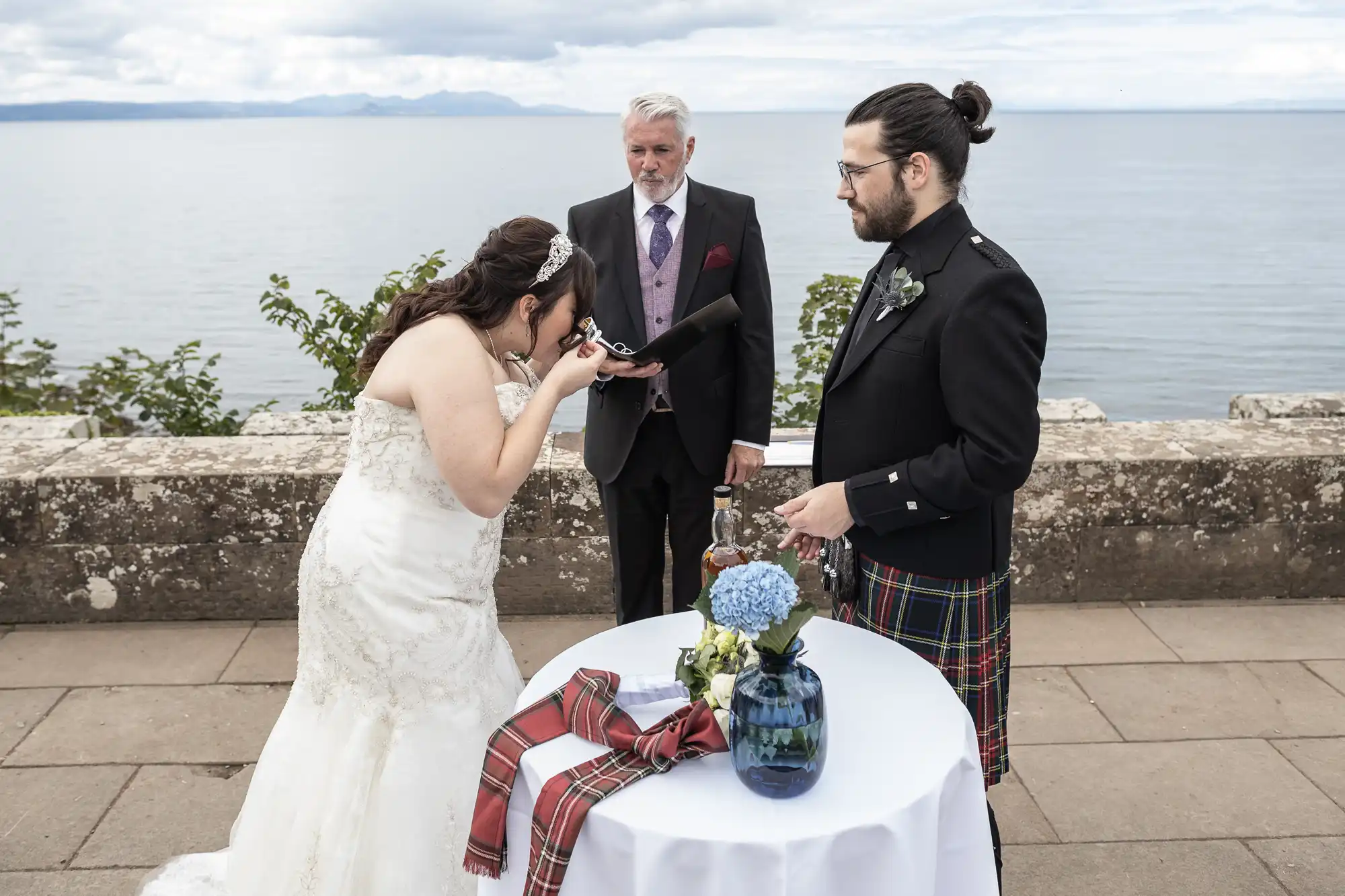 A bride and groom participate in a wedding ceremony by a body of water, with the bride kissing a ceremonial object. An officiant stands behind them. A table with a floral arrangement and tartan sashes is in front.