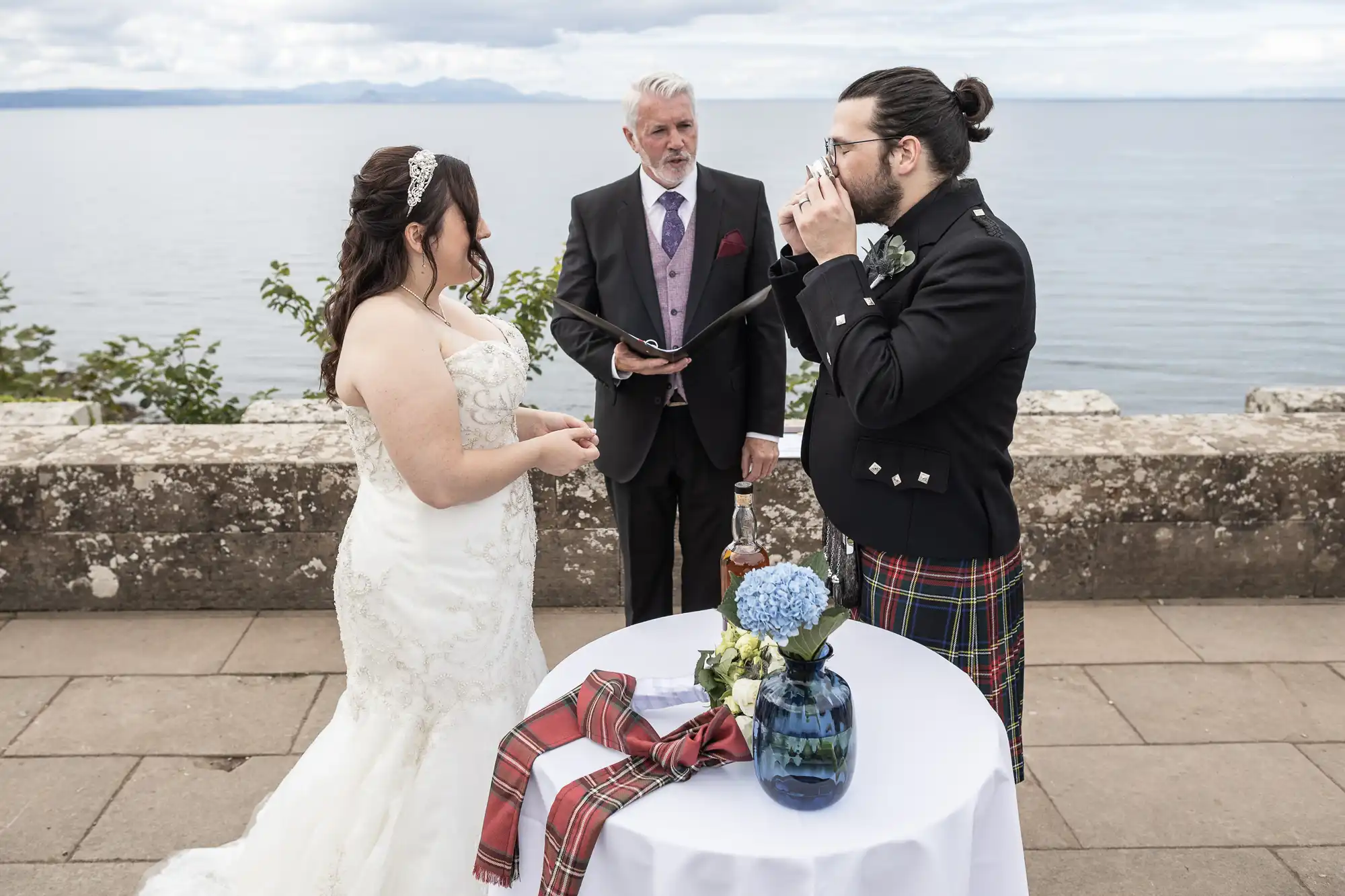 A bride and groom stand on either side of a small table, with an officiant behind them. The groom, dressed in a kilt, drinks from a cup. The bride looks on, with a scenic ocean backdrop.