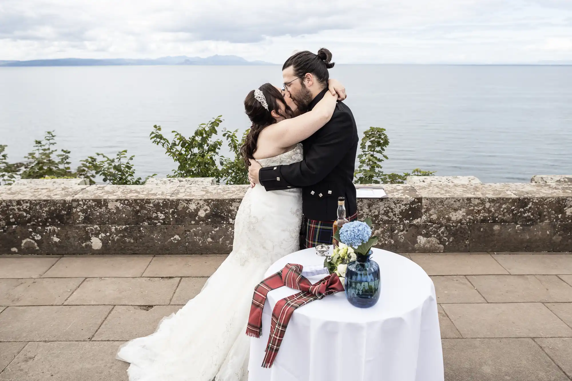 A bride and groom share an embrace near a stone wall overlooking the ocean, with a small table set with a blue vase, flowers, and a tartan cloth in the foreground.