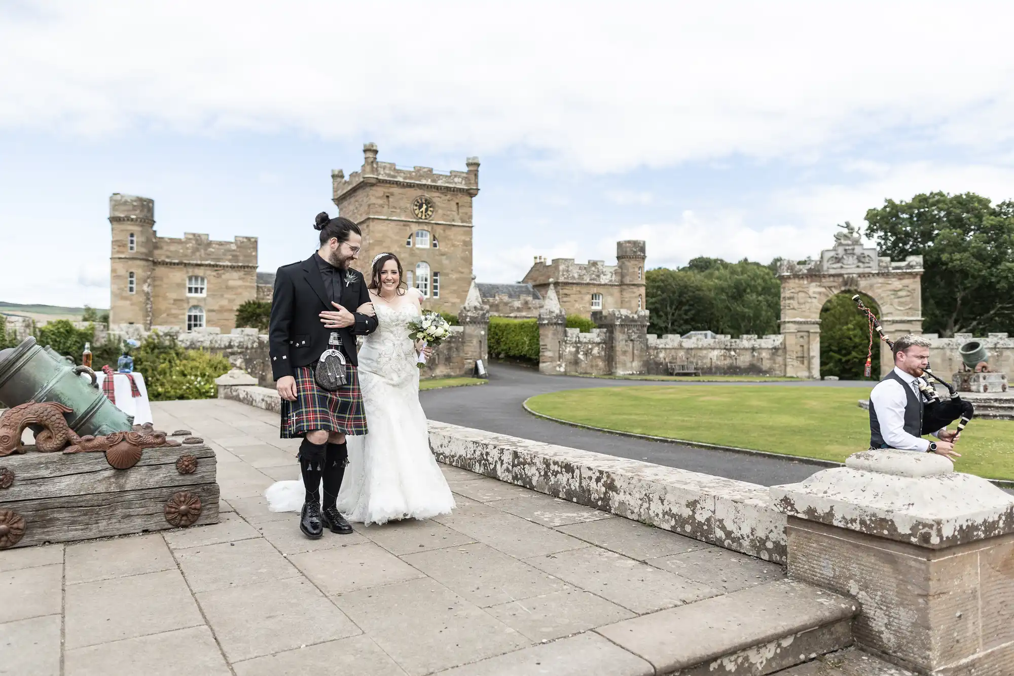 A bride in a white dress and a groom in a kilt walk arm in arm on a stone terrace. A bagpiper plays nearby. A historic stone building and a grassy area can be seen in the background.