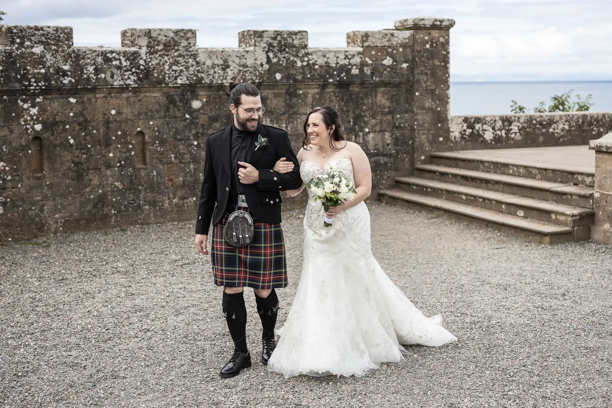 A bride in a white wedding dress and a groom in a traditional kilt walk arm in arm outside a stone building with a view of the sea in the background. Both are smiling and looking at each other.