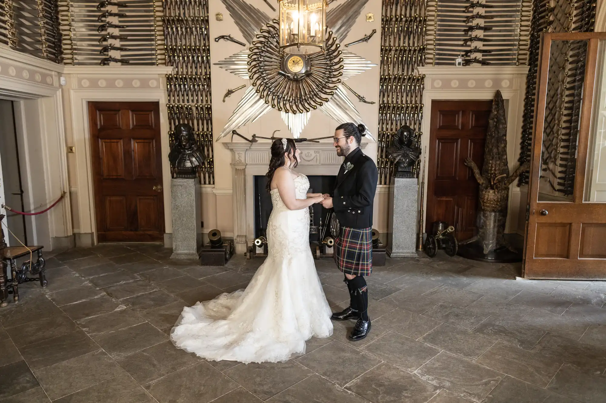 A couple stands holding hands in a room with ornate décor, including an elaborate display of weapons on the wall and a chandelier. The bride wears a white gown, and the groom wears a kilt and jacket.