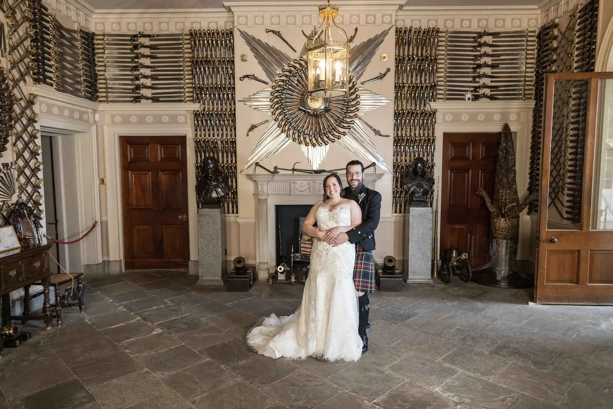 A couple wearing wedding attire stands in an ornate room decorated with swords and shields, posing in front of a fireplace under a large circular ornament of weapons.