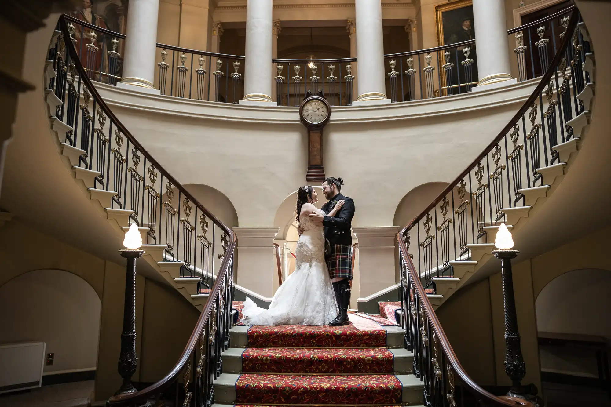 A couple embraces on a grand staircase inside a historic building with columns, ornate railings, and a clock above the landing. The bride wears a white gown, and the groom wears a kilt.