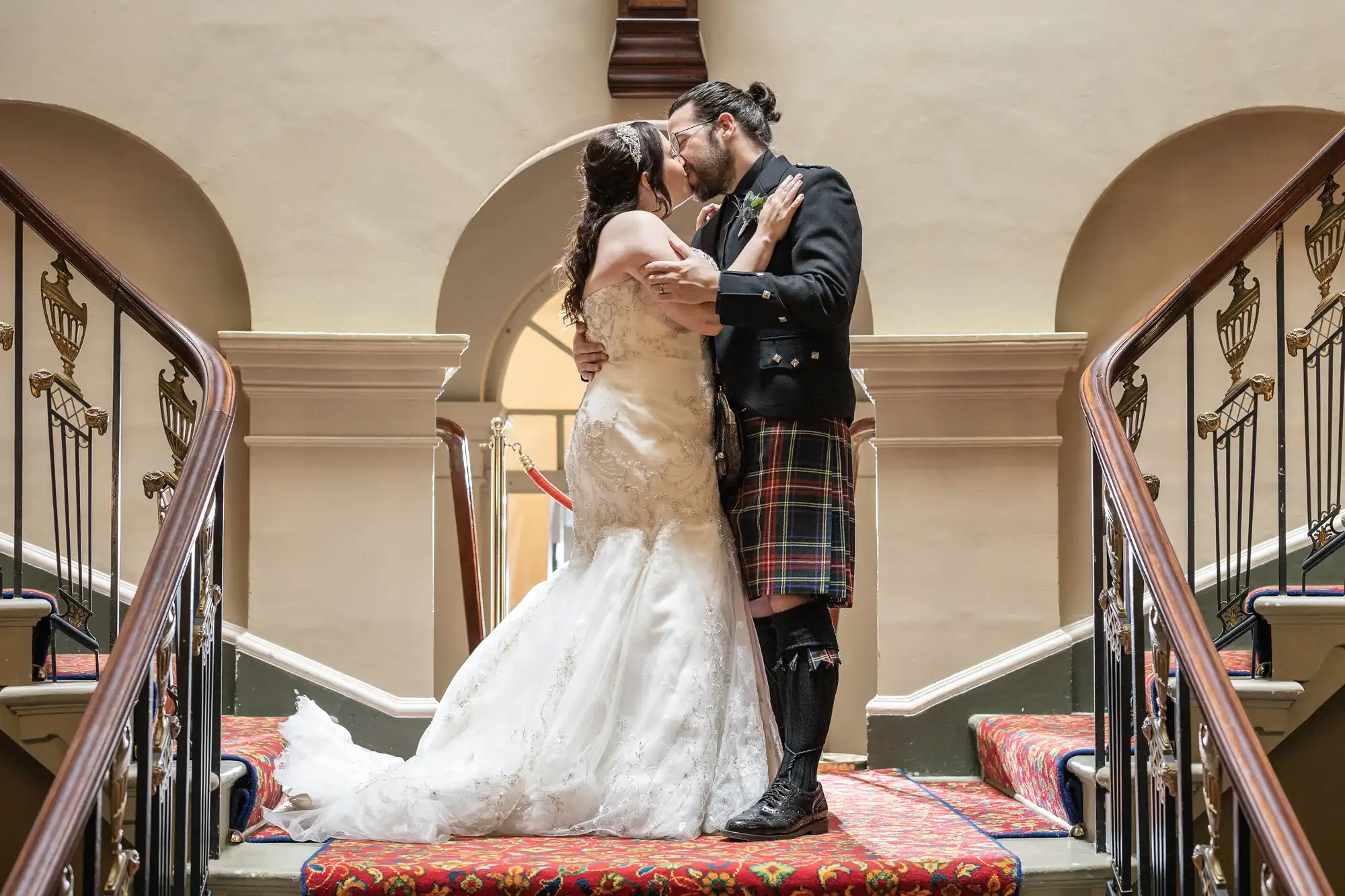 A couple kisses at the top of a staircase, with the bride in a white gown and the groom in a traditional Scottish outfit.