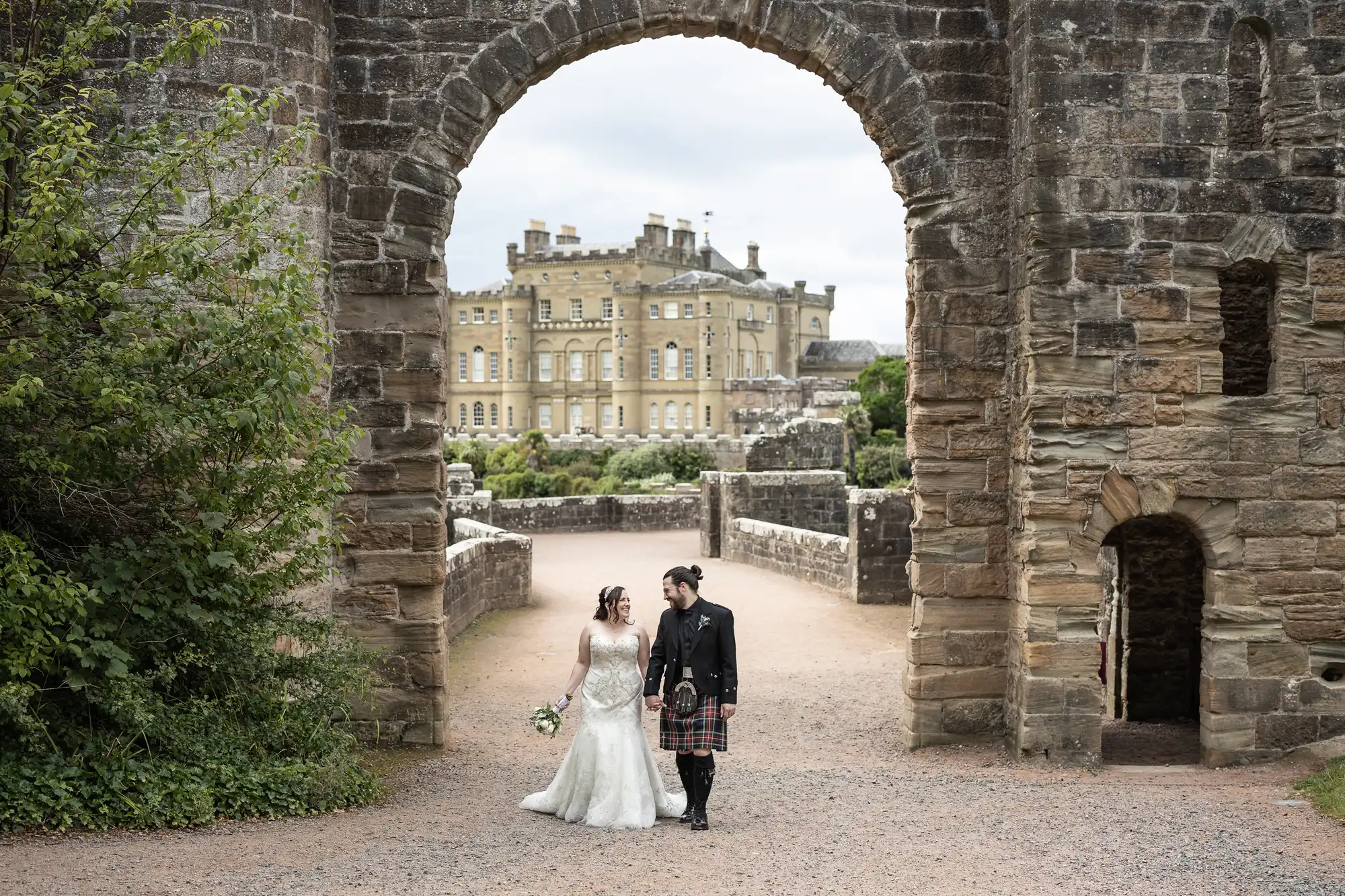 A bride and groom, dressed in a wedding gown and traditional Scottish attire respectively, hold hands while walking through an ancient stone archway with a castle in the background.