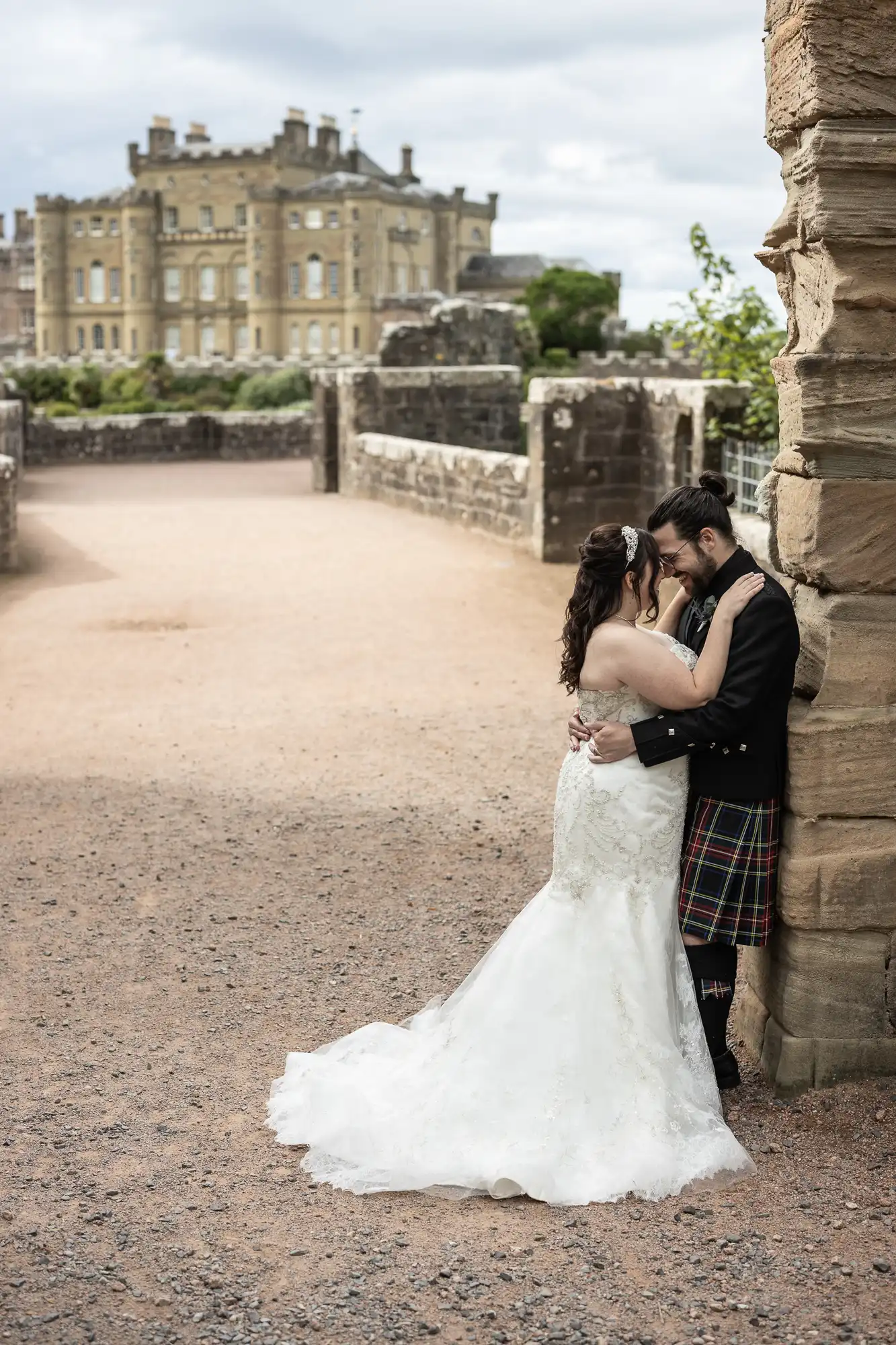 A bride in a white gown and a groom in a kilt embrace beside stone walls, with a large castle in the background.