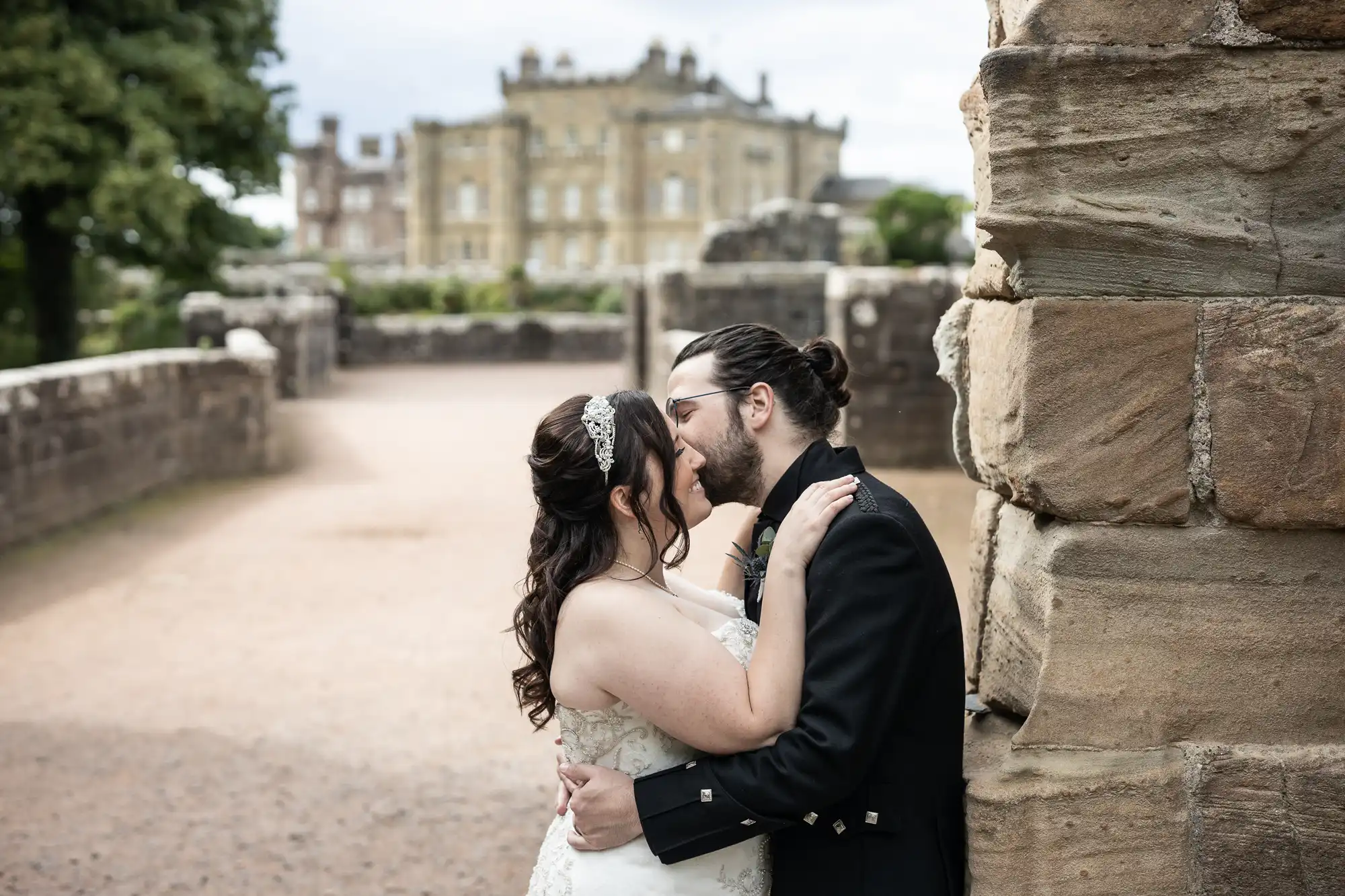 A bride and groom dressed in formal attire are embracing and kissing beside a stone wall with a large mansion blurred in the background.