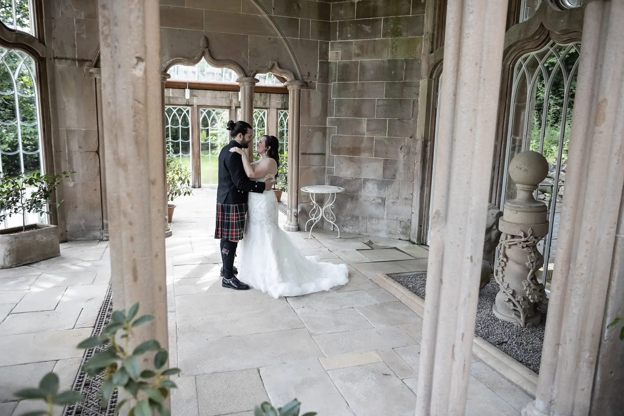 A bride and groom embrace in a stone pavilion with arched windows. The groom wears a kilt and the bride is in a white dress.