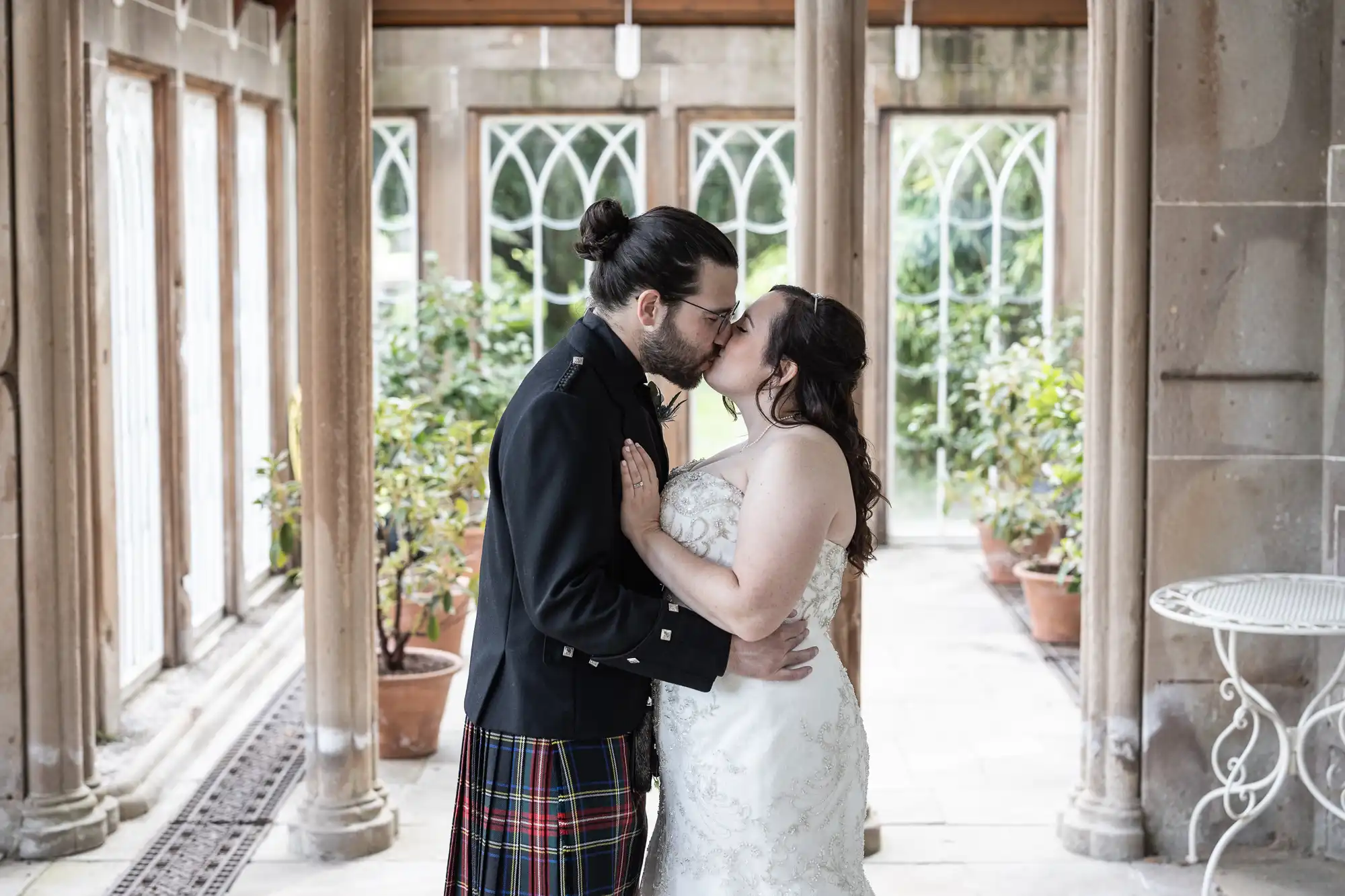 A couple, dressed in wedding attire with the man in a kilt and the woman in a white gown, kiss in a columned walkway with windows and potted plants in the background.