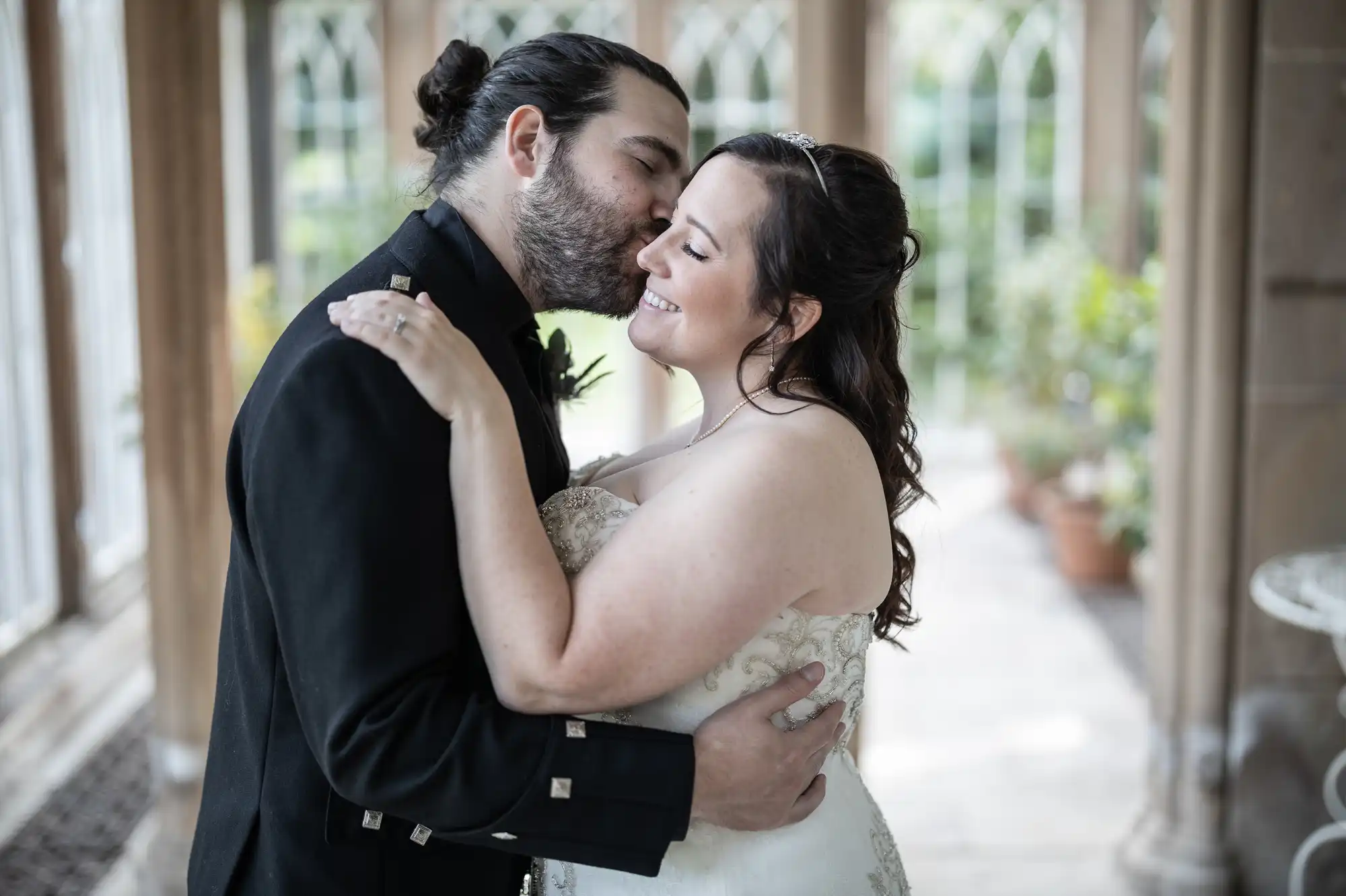 A couple embraces and shares a kiss inside a sunlit room with large arched windows.