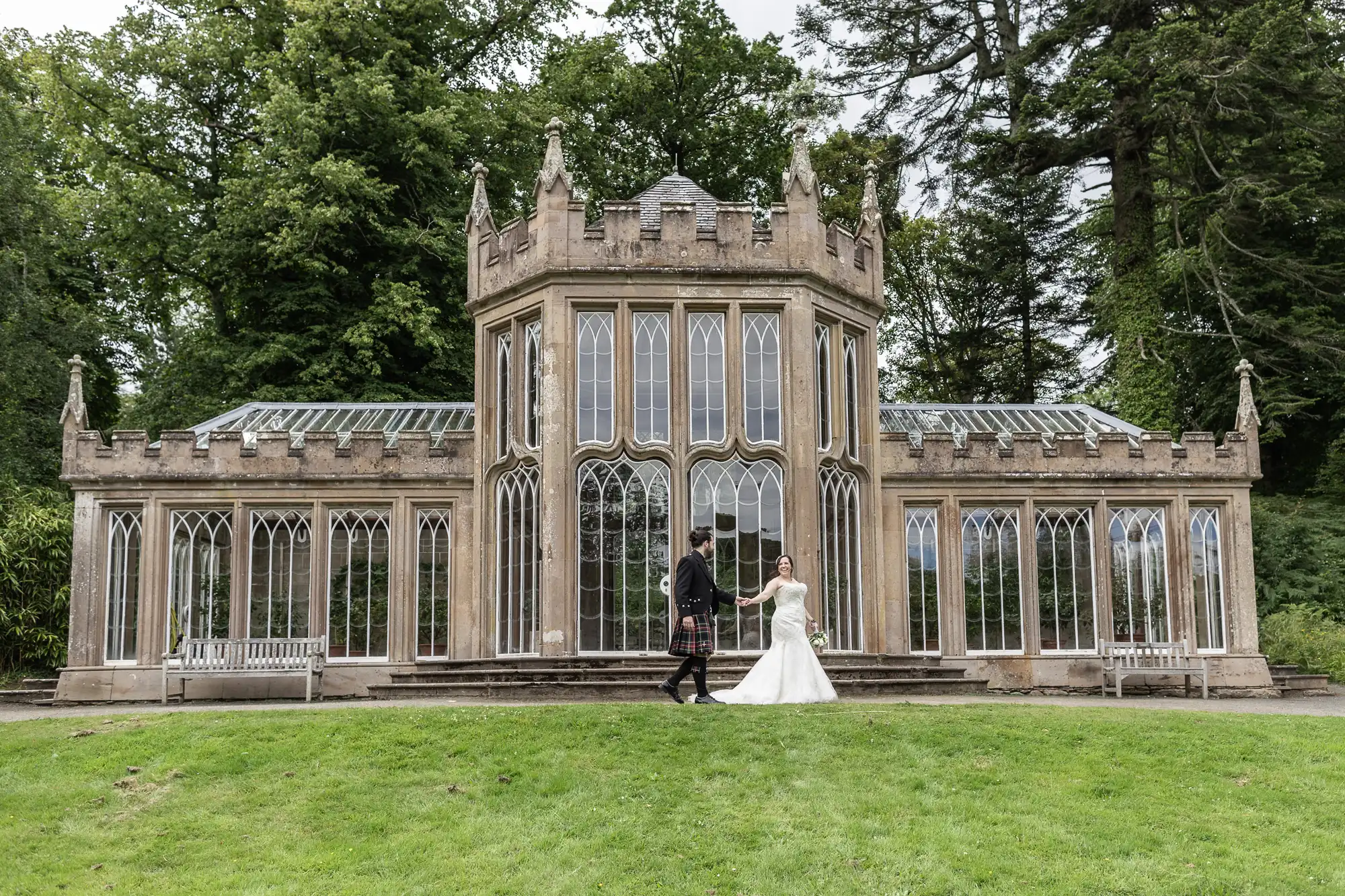 A couple, dressed in formal attire, hold hands in front of a Gothic-style conservatory with tall, arched windows and surrounded by lush greenery.