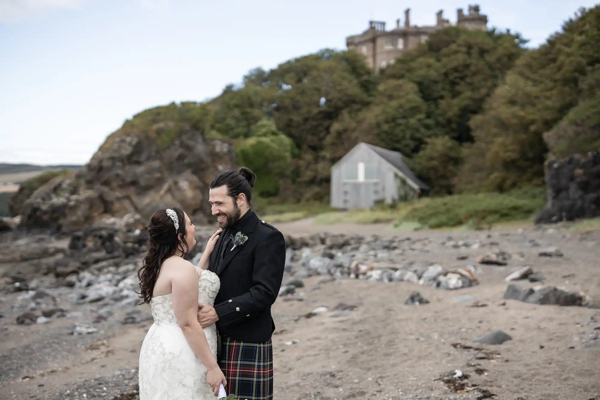 A bride and groom, with the groom wearing a kilt, stand on a rocky beach in front of a hill with trees and buildings in the background.