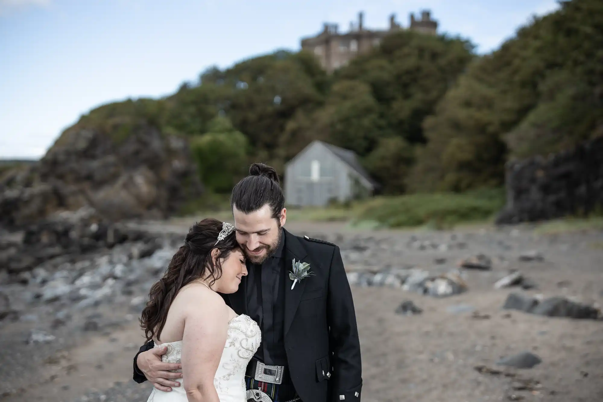 A newlywed couple embraces on a rocky beach with greenery and a large house in the background. The groom wears a dark suit while the bride is in a strapless white dress.