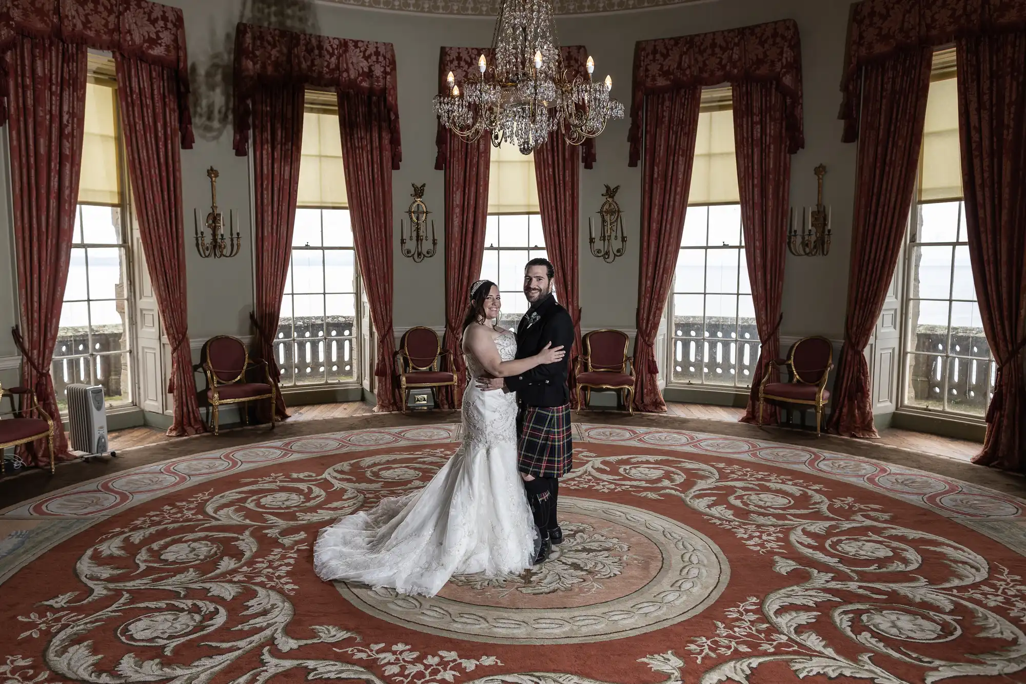 A bride in a white gown and a groom in a kilt stand together, posing in an ornate room with red curtains, a chandelier, and a large circular decorative carpet.