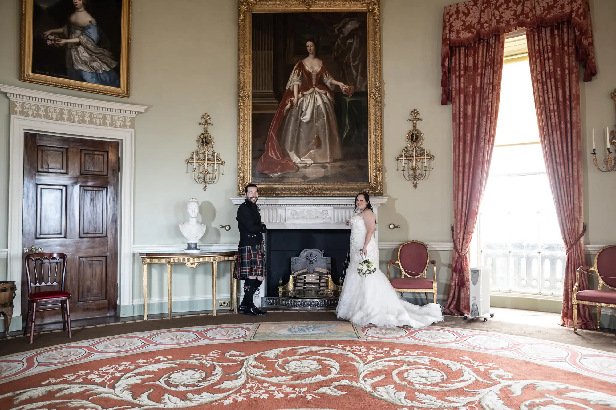 A couple in formal attire stands in an opulent room with a large portrait over the fireplace. The man is in a kilt; the woman in a wedding dress holds a bouquet. The room features ornate decor and red curtains.