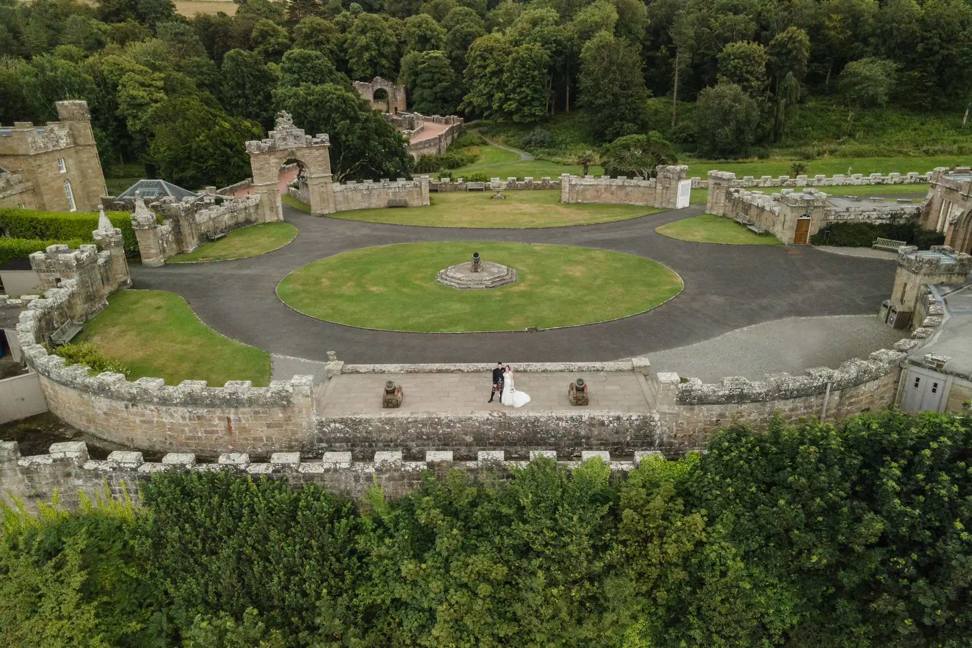 Aerial view of a stone castle with surrounding greenery, featuring a circular driveway and a statue at the center. A couple in wedding attire stands on the castle wall.