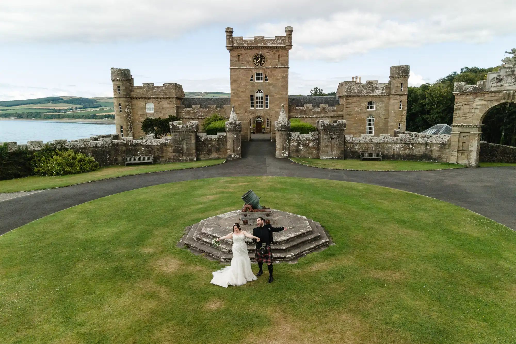 A bride in a white dress and a groom in a kilt hold hands on a circular stone platform, with a historic castle in the background, surrounded by green lawns.