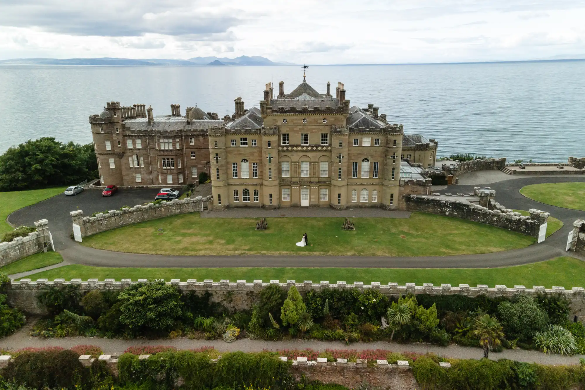 A large historic building sits by the sea, surrounded by lush greenery. A couple stands on the lawn, dressed in formal attire.