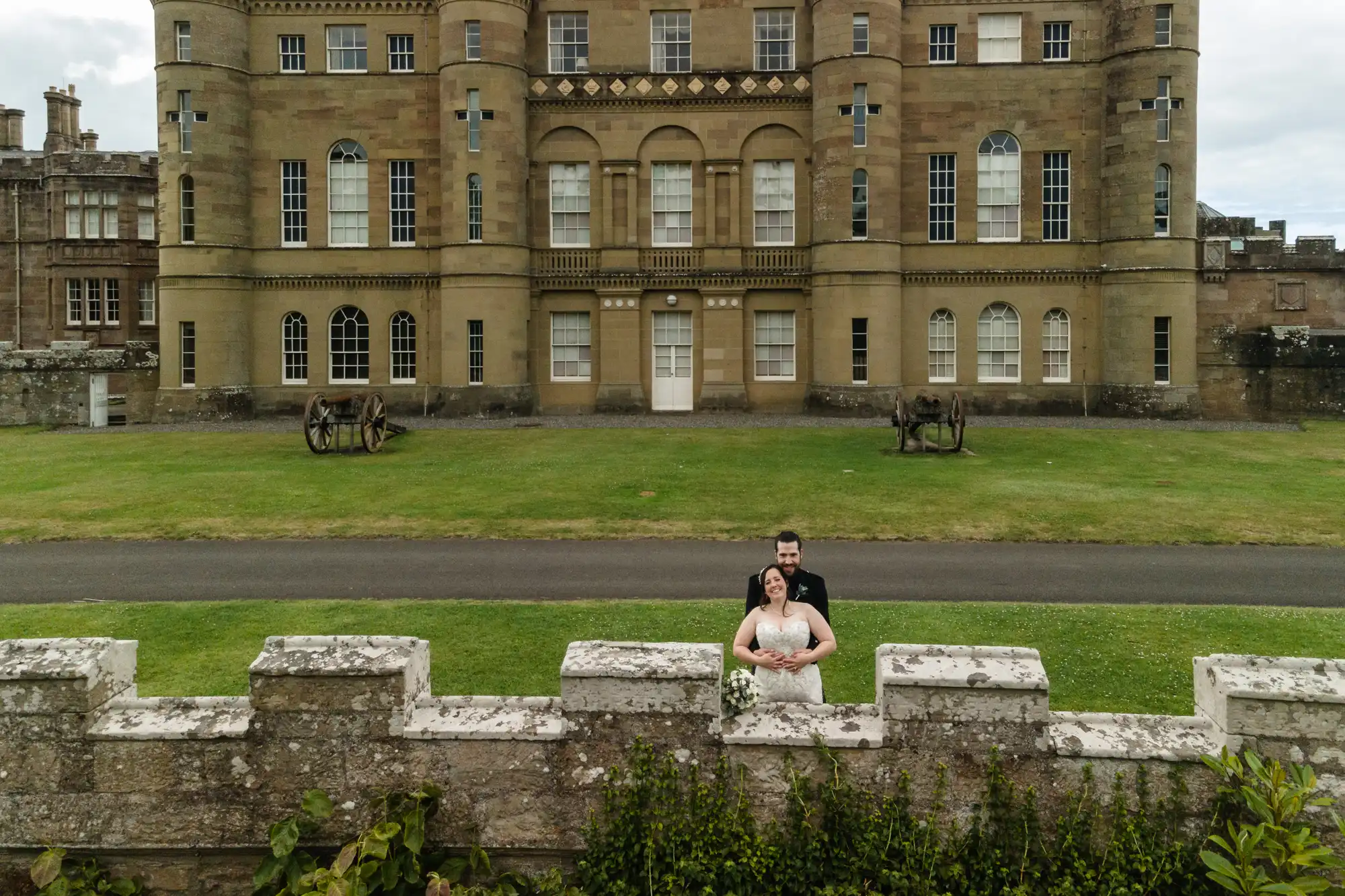 A newlywed couple stands in front of a large historic stone building with green lawns and a low stone wall in the foreground.