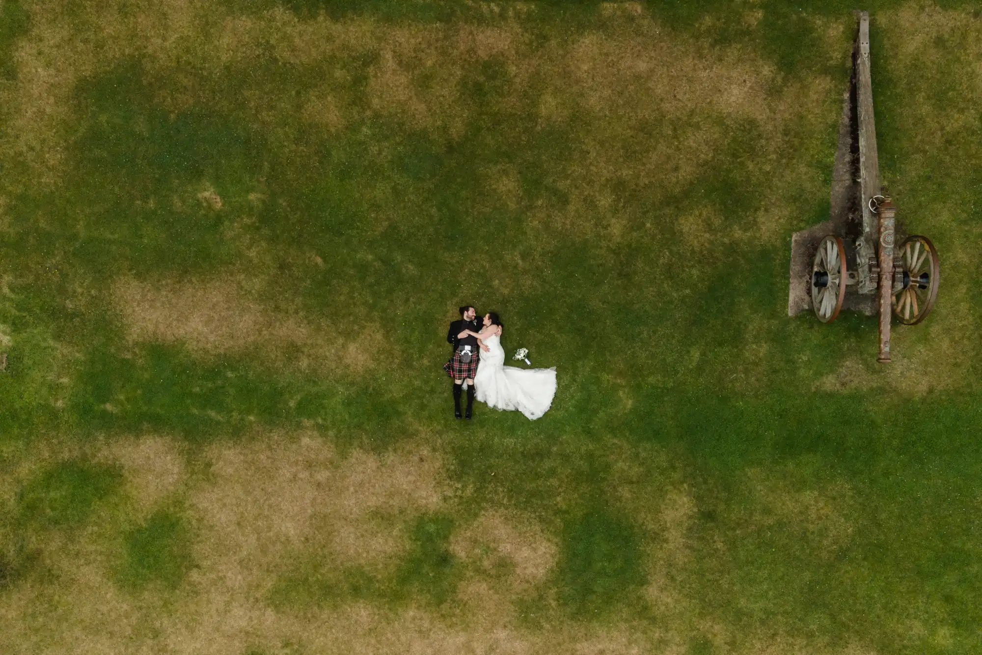 Aerial view of a couple lying on the grass. The woman in a white dress and the man in a kilt are side by side. An antique cannon is located to their right.