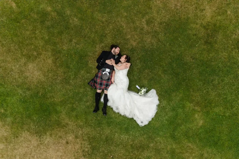 Culzean Castle wedding photos: Aerial view of a couple in wedding attire lying on a grassy field. The groom wears a kilt, and the bride is in a white wedding dress holding a bouquet.