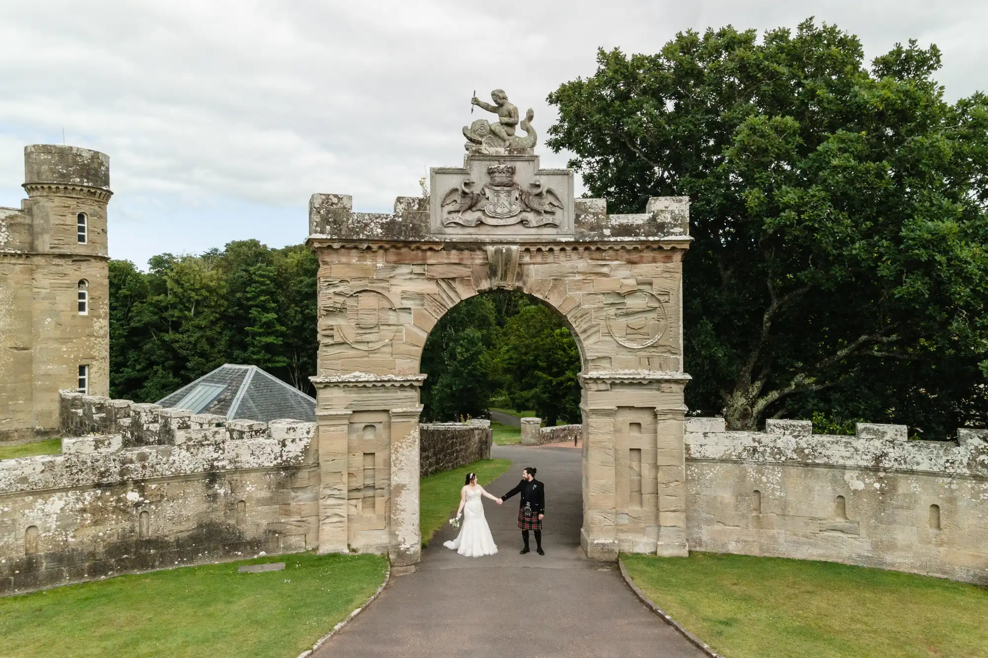 A couple in wedding attire stands holding hands under a historic stone archway with greenery in the background.