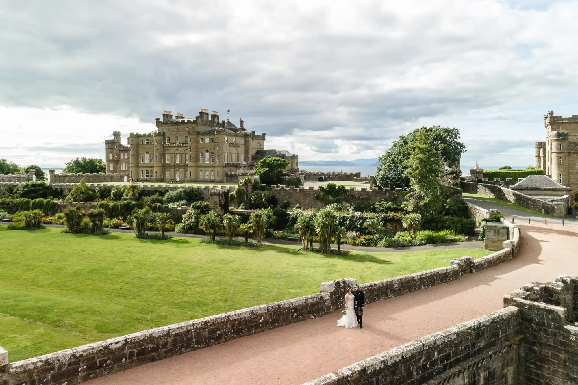 A couple dressed in wedding attire walk along a paved path near an expansive castle with lush greenery and gardens surrounding it under a cloudy sky.