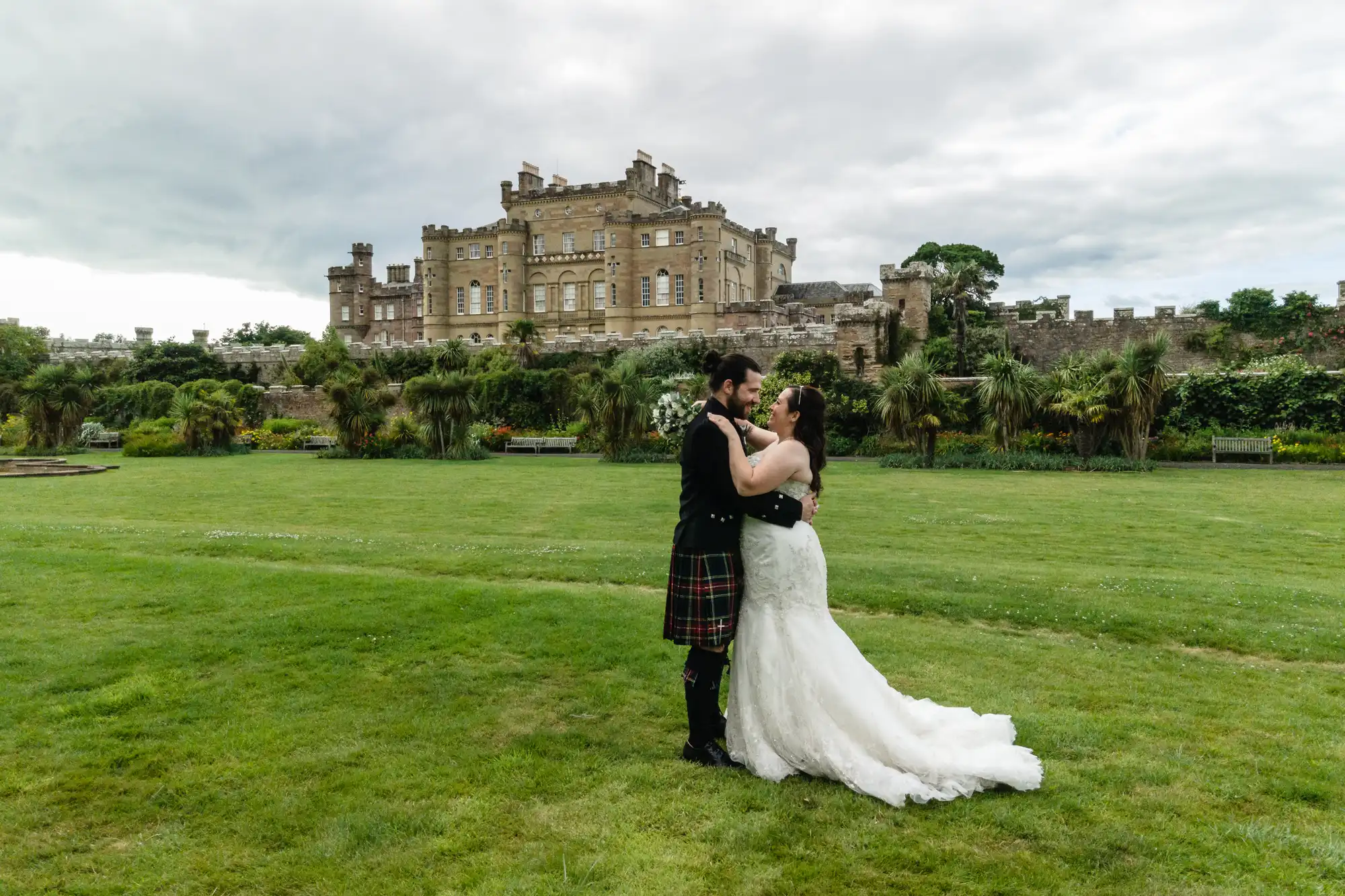 A couple dressed in wedding attire embrace on a grassy lawn with an old castle in the background.