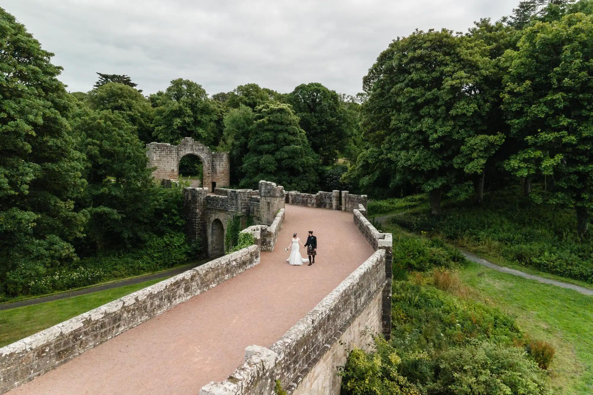 A bride and groom walk hand in hand across a historic stone bridge surrounded by lush greenery.