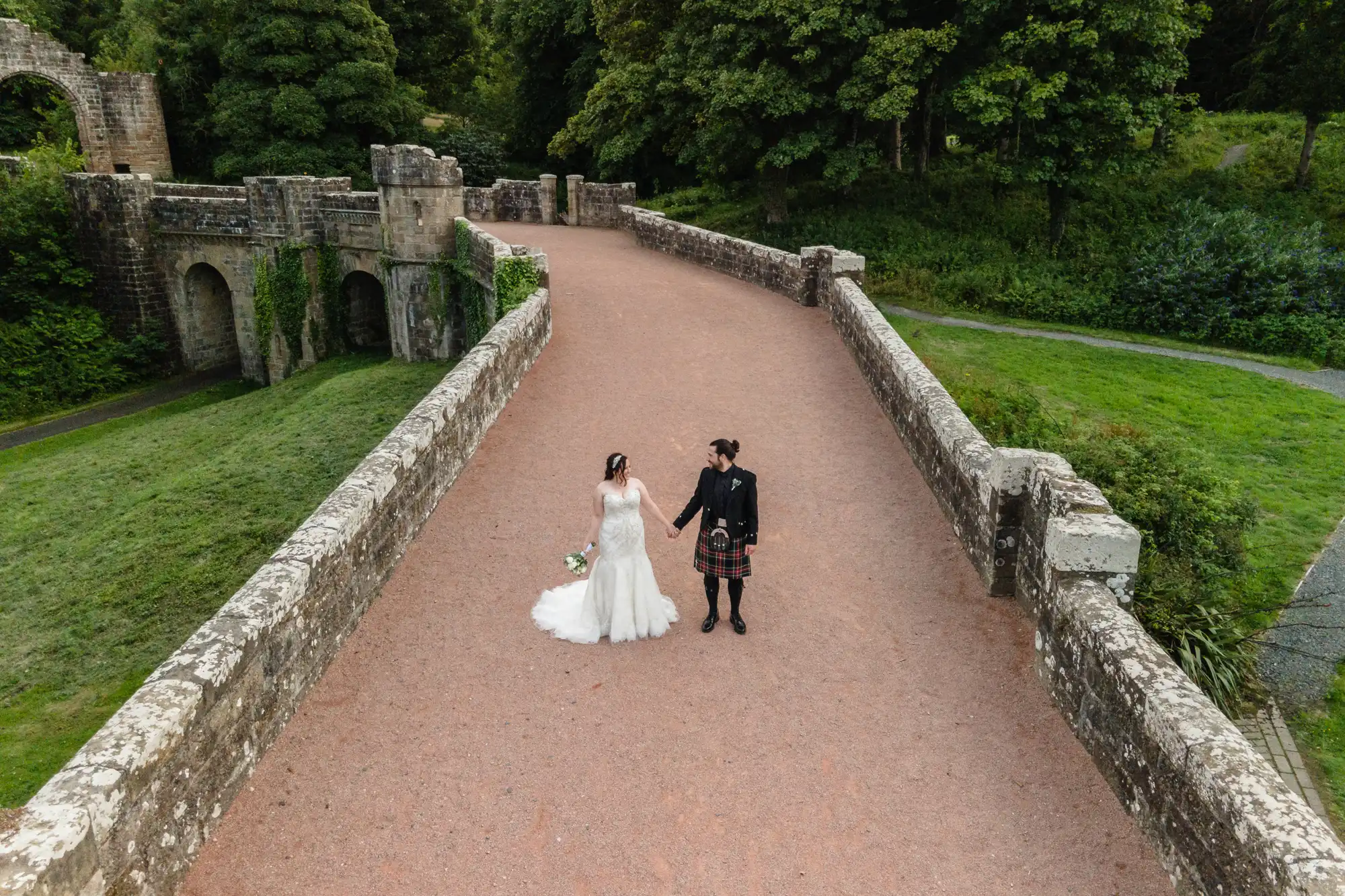 A bride and groom stand on a stone path holding hands. The groom is dressed in traditional Scottish attire and the bride wears a white gown. Trees and stone structures are visible in the background.