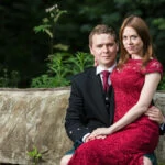 bride wearing red lace dress sits on the knee of her husband wearing a kilt outfit