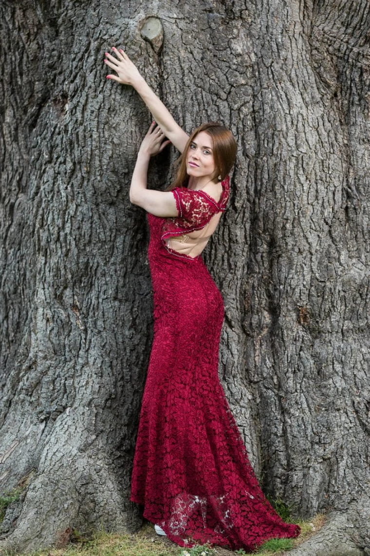 bride wearing a red lace dress poses in front of the large tree on the castle lawn