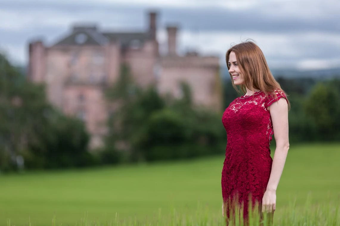 bride wearing a red lace dress standing in the field with the castle in the background