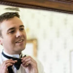 groom adjusts his bow tie in front of a mirror